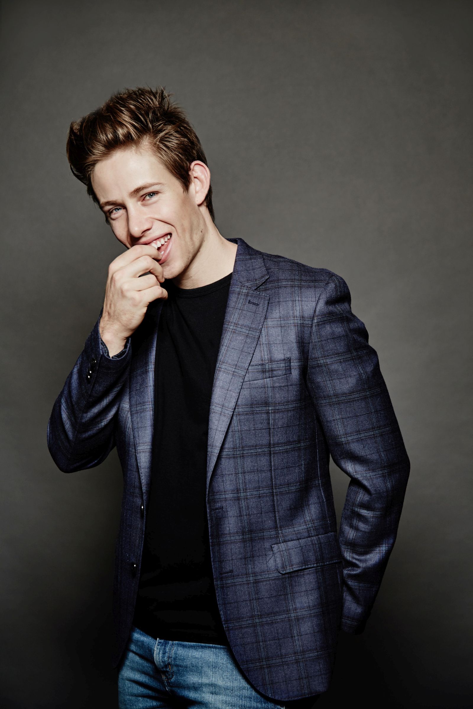 Studio portrait of young man smiling, hand at mouth, blue plaid blazer and jeans