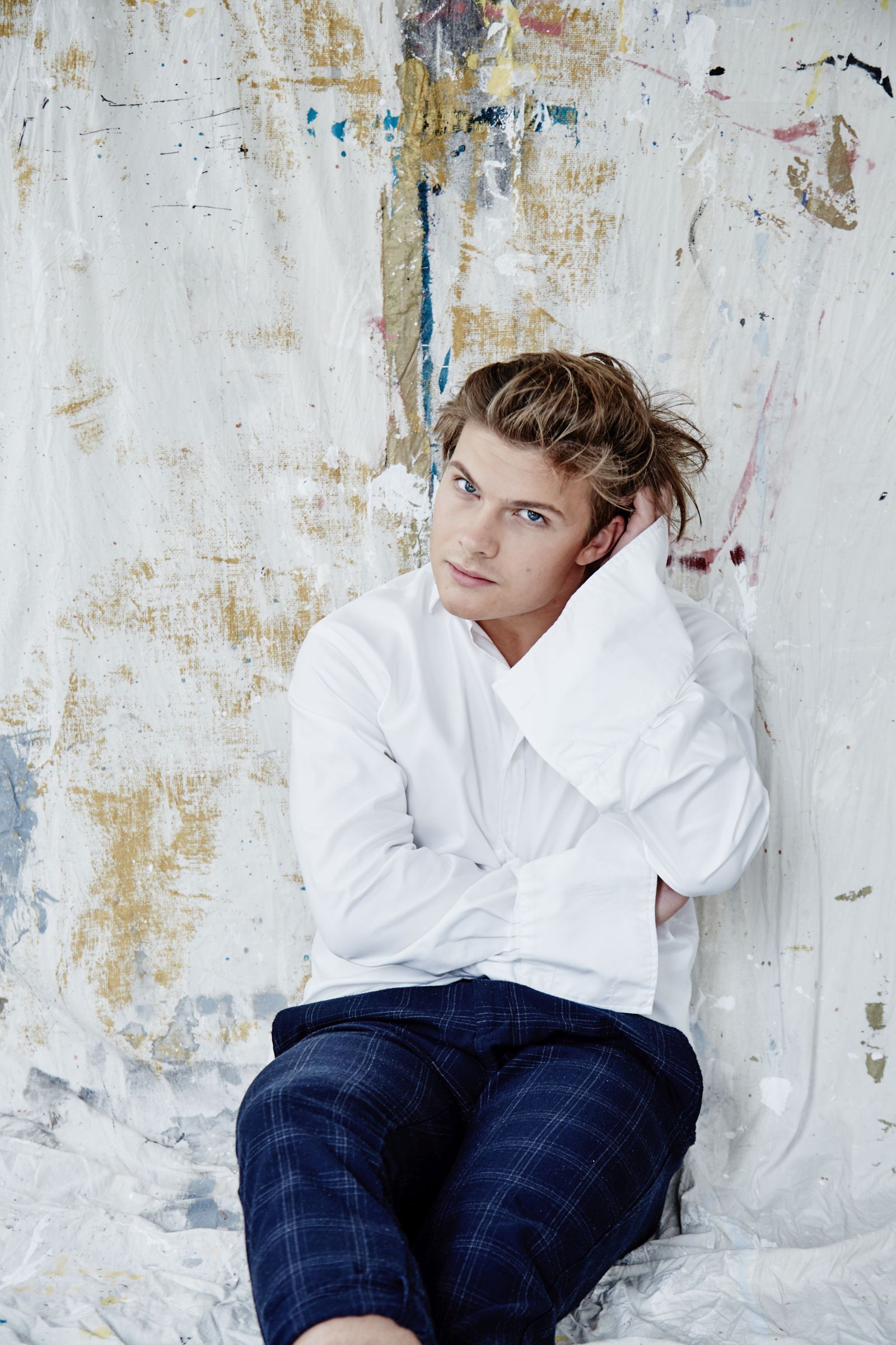 Editorial portrait of person in white shirt, hand in hair, against paint-splattered canvas.