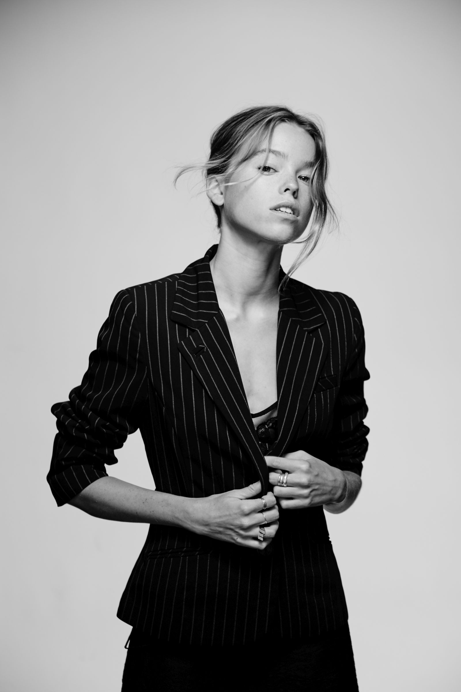 Black-and-white editorial portrait of model in pinstripe blazer, adjusting lapels, rings visible.