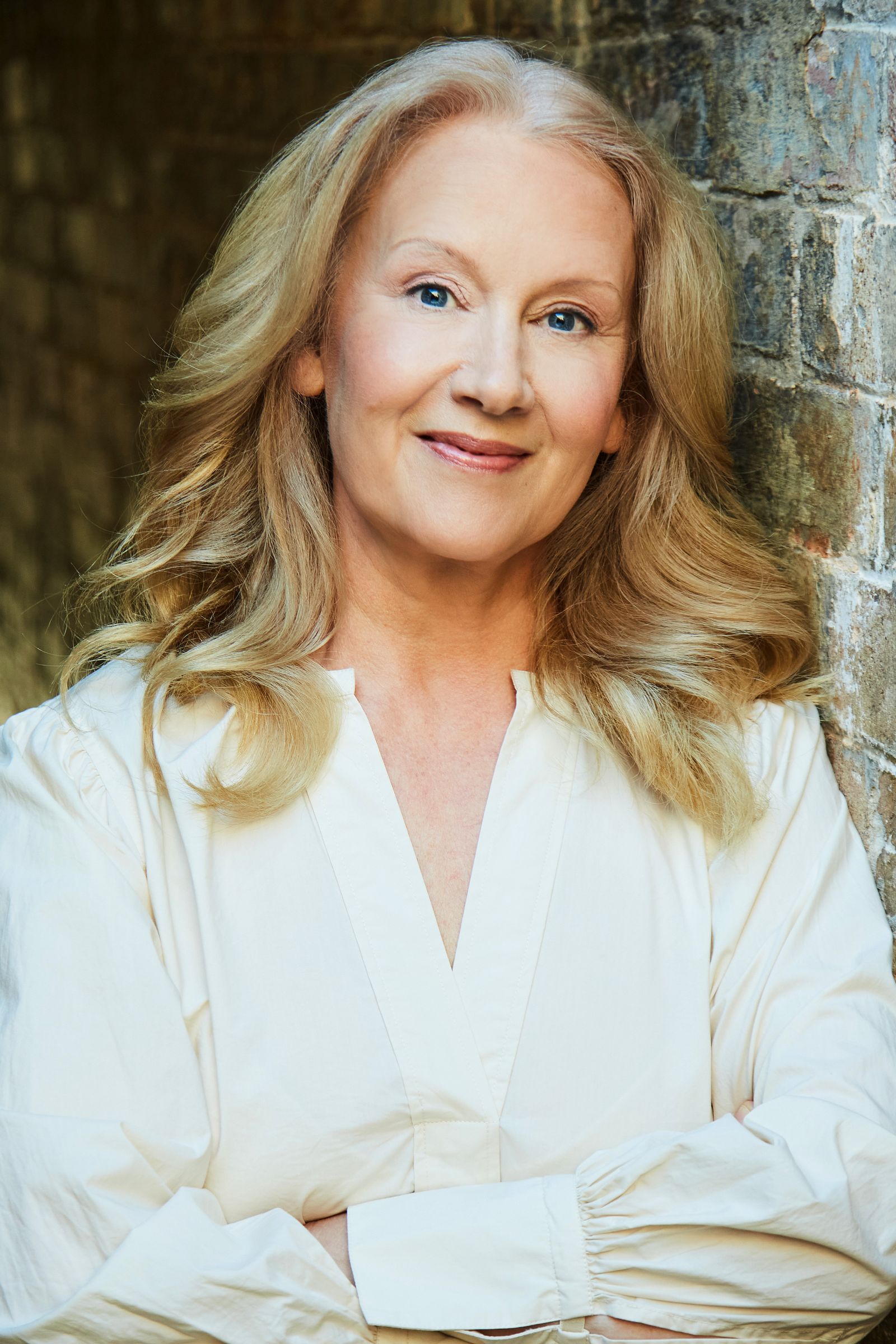 Professional headshot of smiling woman with long blonde hair, white blouse, leaning on brick wall.