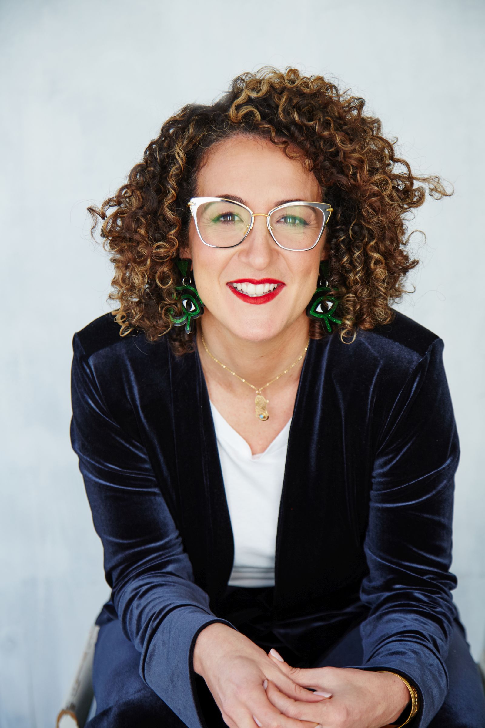 Studio portrait: smiling person with curly hair, glasses, red lipstick, green earrings, velvet blazer.