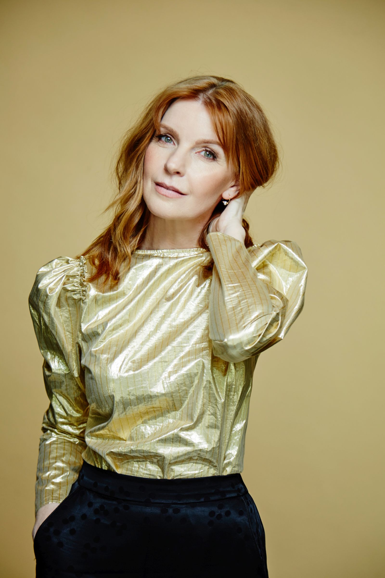 Editorial studio portrait of red-haired woman in metallic gold blouse, beige background.