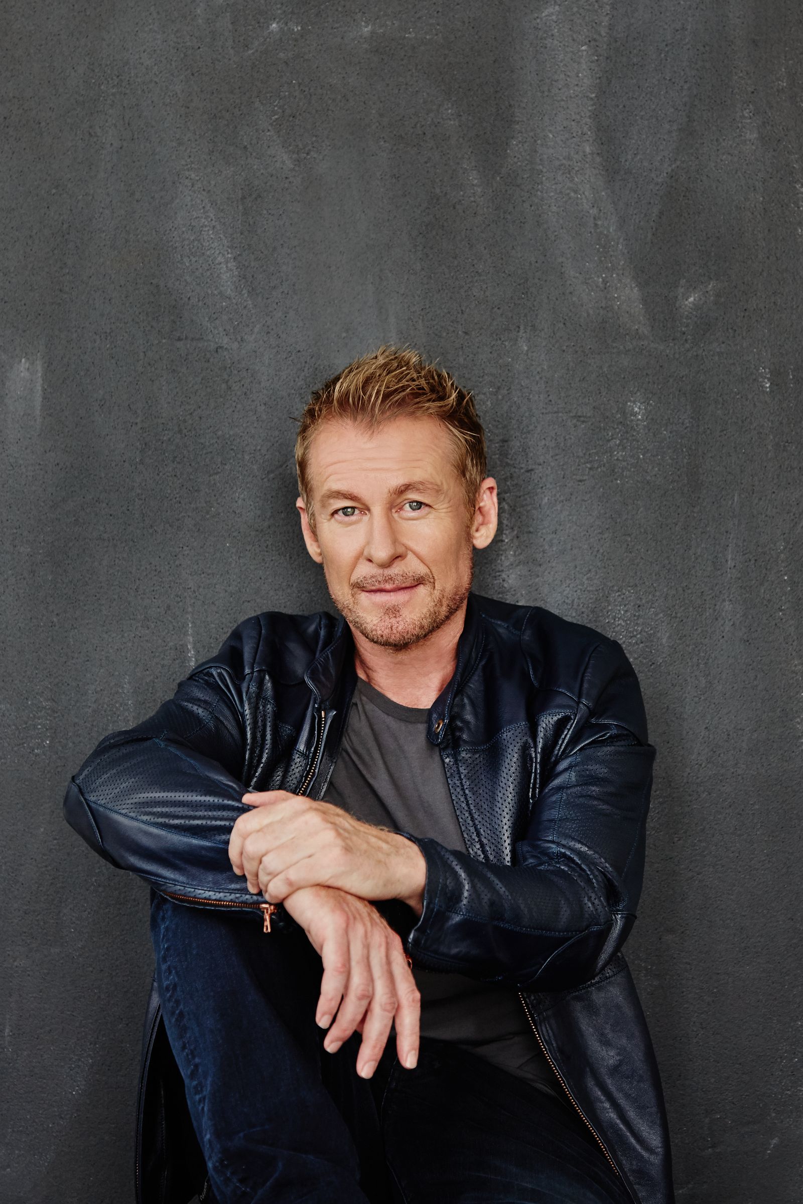 Casual studio portrait of blond man in navy leather jacket, seated against dark wall.
