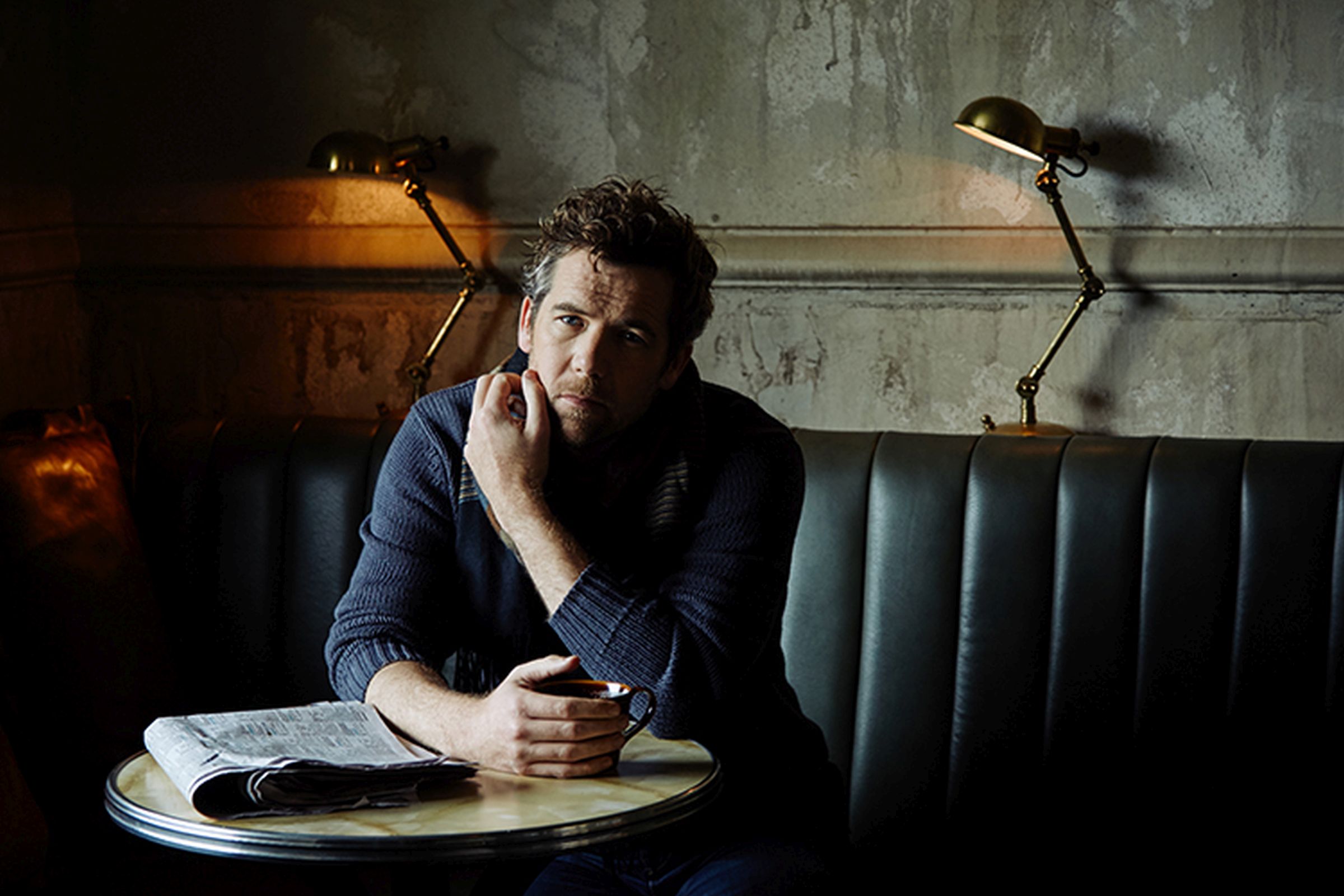 Pensive man in café booth holding coffee, newspaper on table, brass wall lamps.