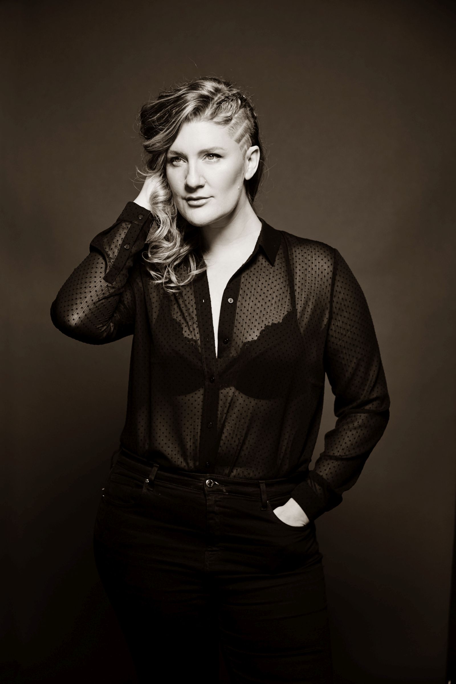 Monochrome studio portrait, person with side-shaved hair, sheer dotted blouse, hand in pocket.