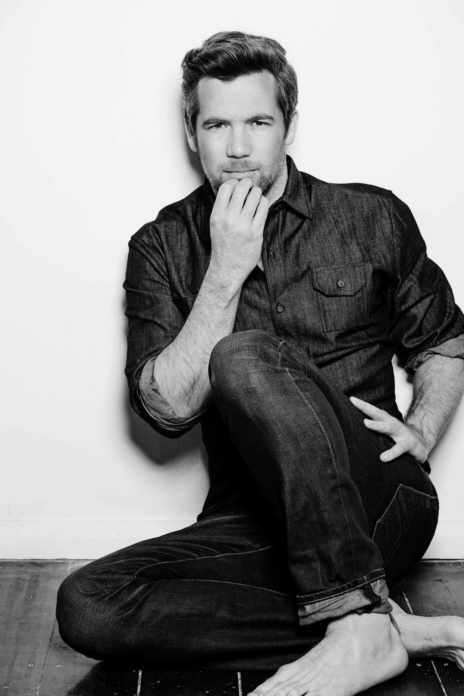 Black and white editorial portrait of barefoot man in denim, seated, hand to chin.