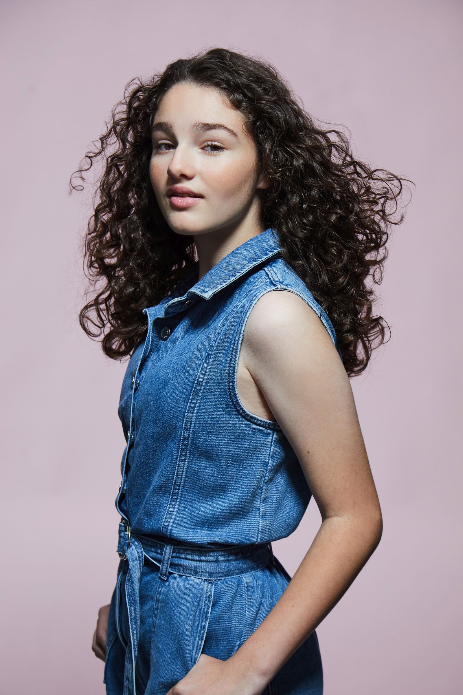 Young woman with curly hair wearing a denim outfit, pink background.