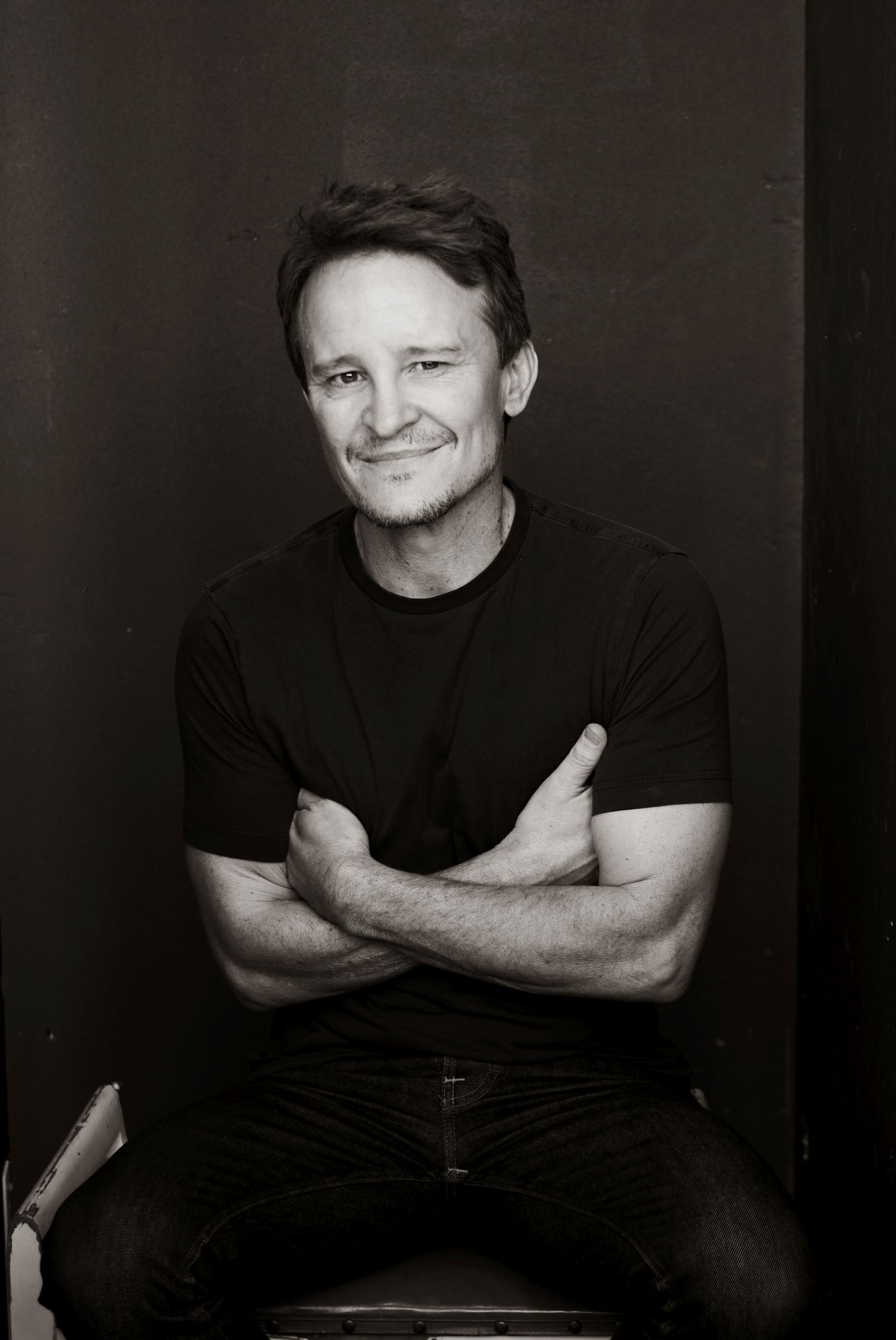 Man in dark shirt sitting with arms crossed, smiling softly.