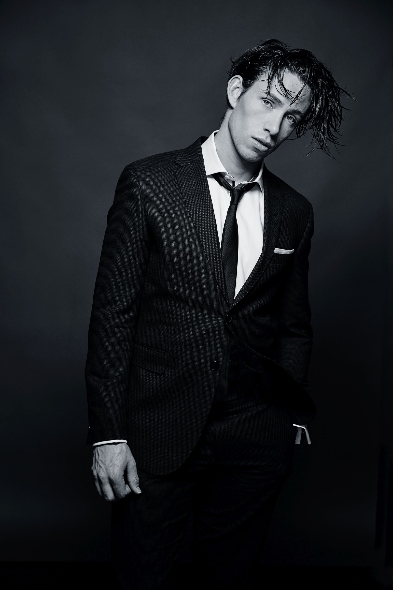 Moody black-and-white studio portrait of person in a suit, wet hair, loosened tie.