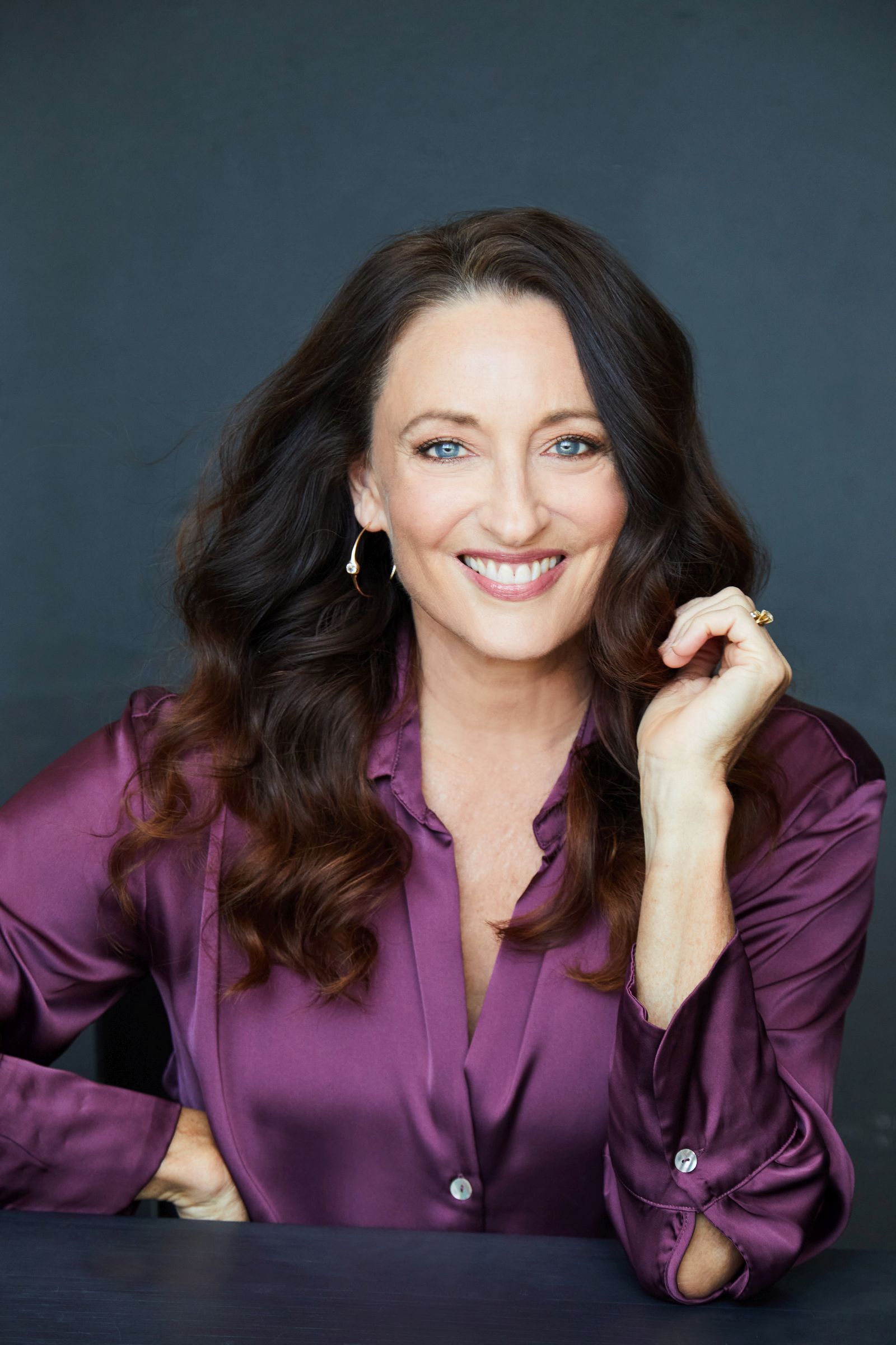 Woman smiling, wearing a purple blouse, with wavy hair against dark background.