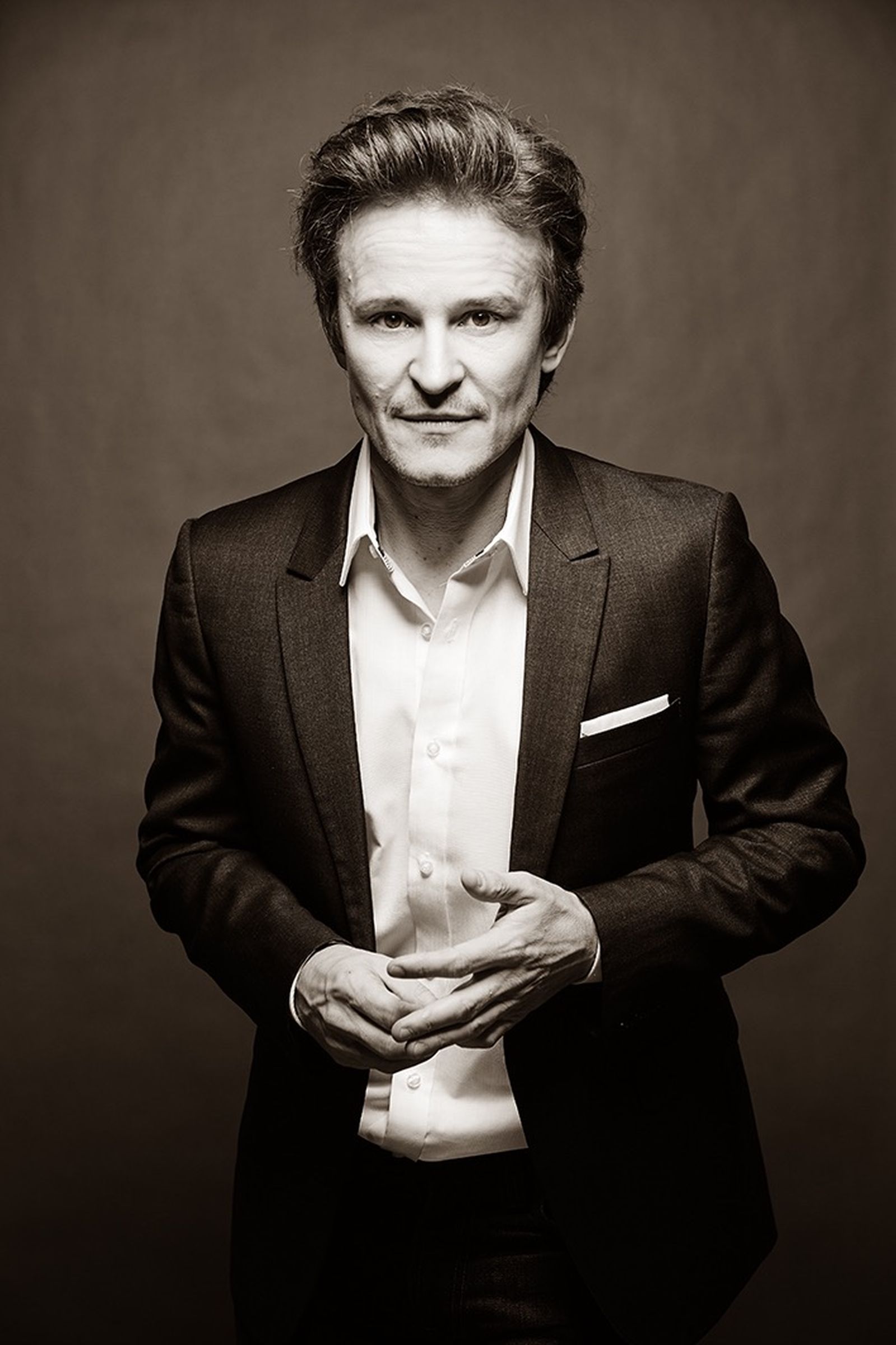 Monochrome editorial portrait of man in suit, direct gaze, hands clasped, studio backdrop.