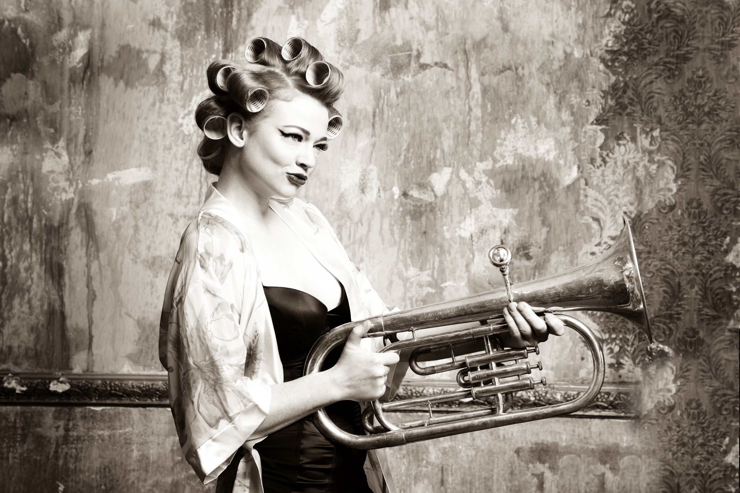 Sepia-toned portrait of person with hair rollers holding a large brass horn, vintage wall.