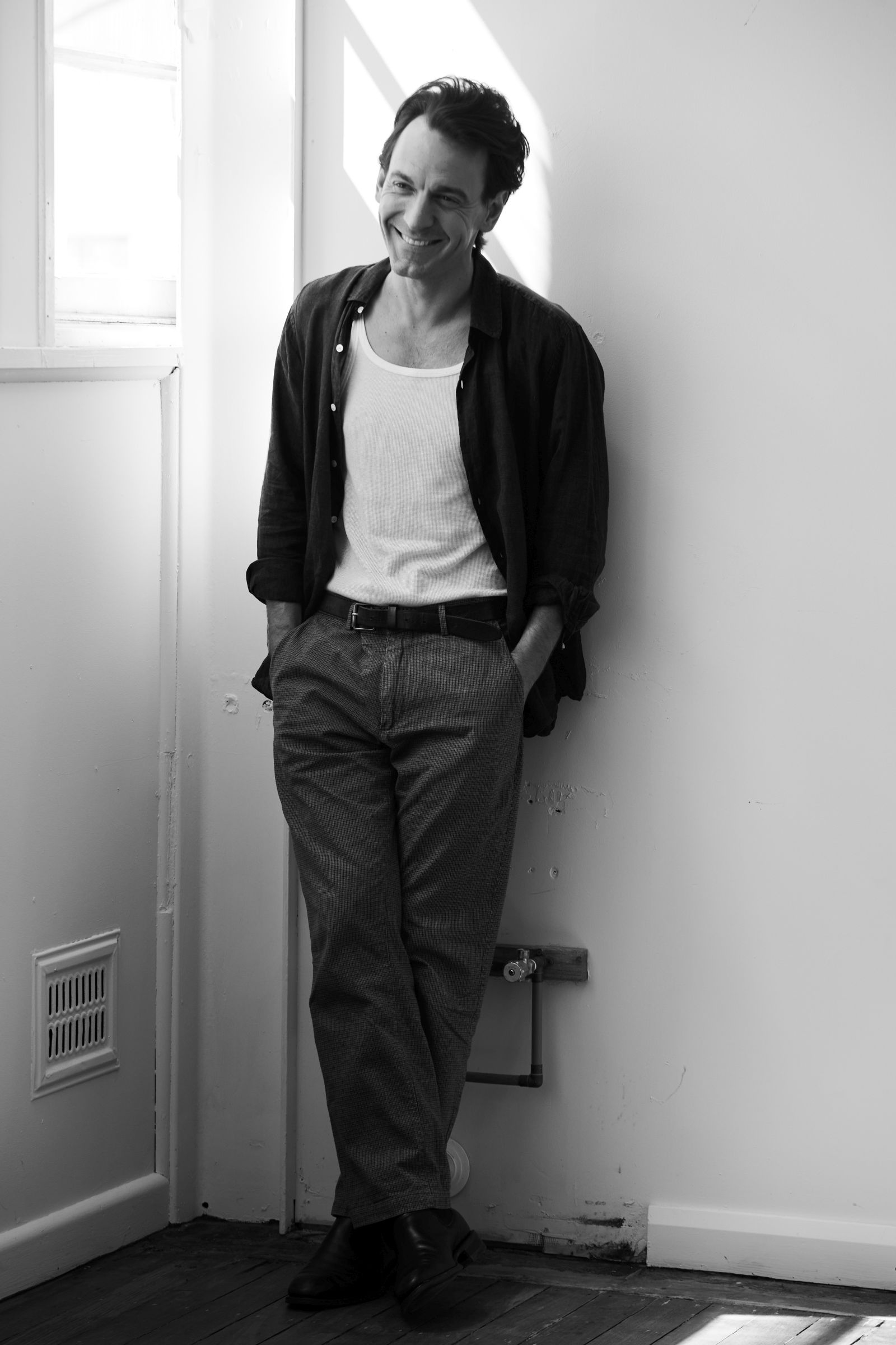 Smiling man in white tank and open shirt leans against sunlit wall.