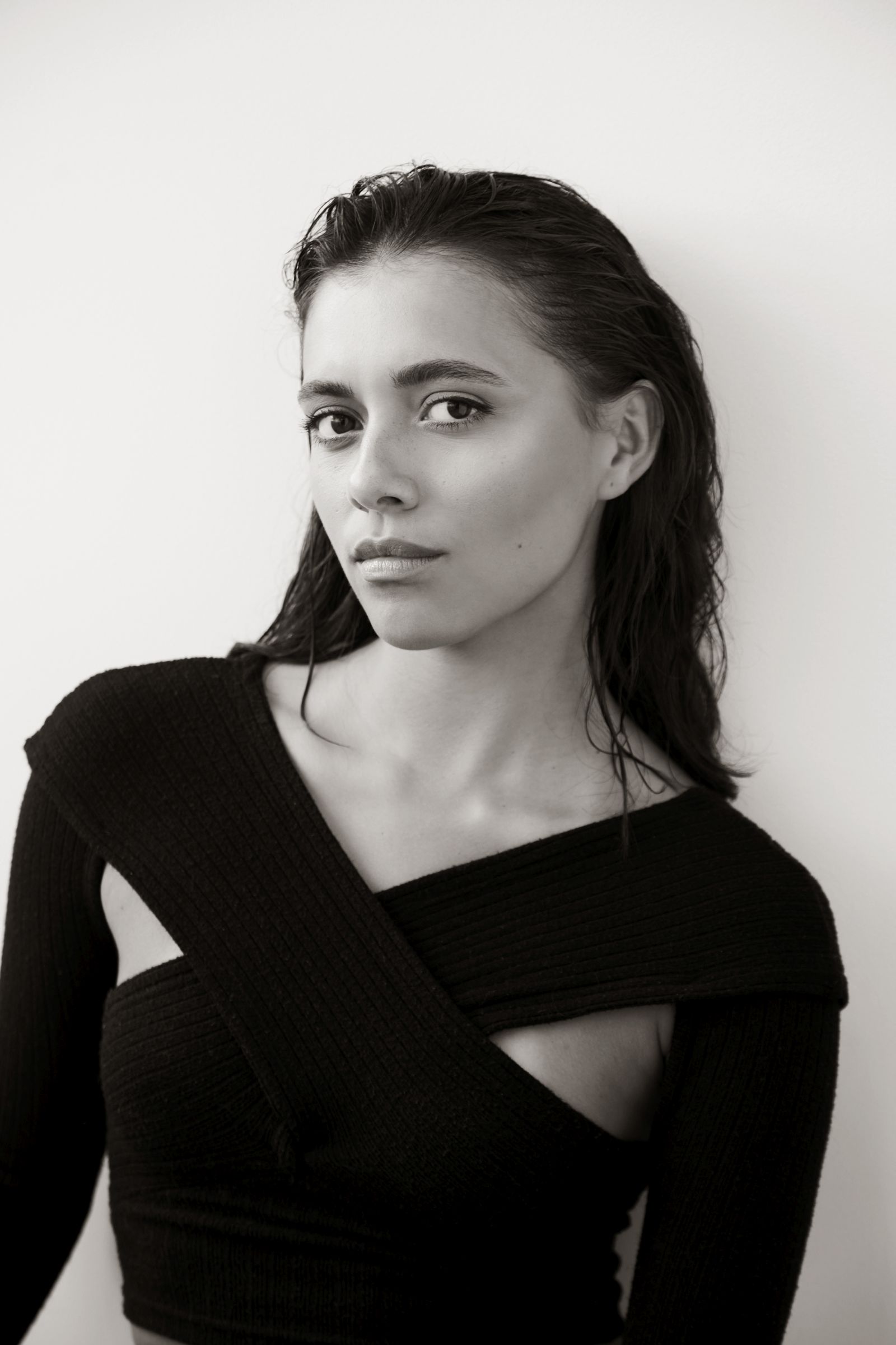 Black-and-white editorial headshot of person with wet hair, asymmetrical black knit top, neutral gaze.