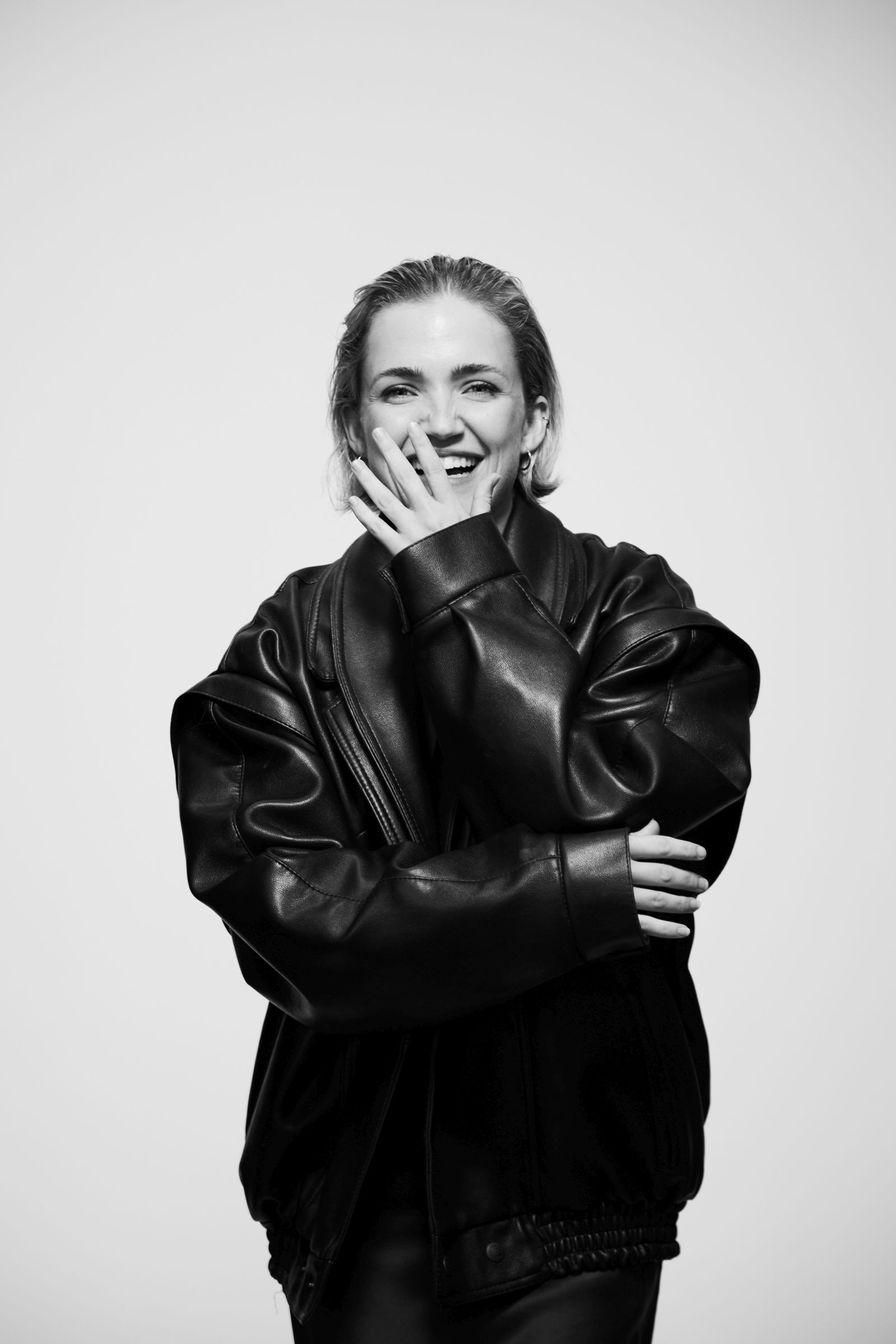 Black-and-white studio portrait: person laughing, hand over mouth, in oversized leather jacket.