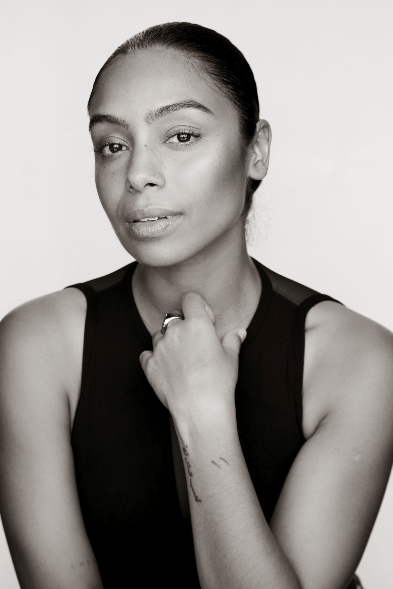 Black and white portrait of a person with a tattoo on their forearm, wearing a sleeveless top.