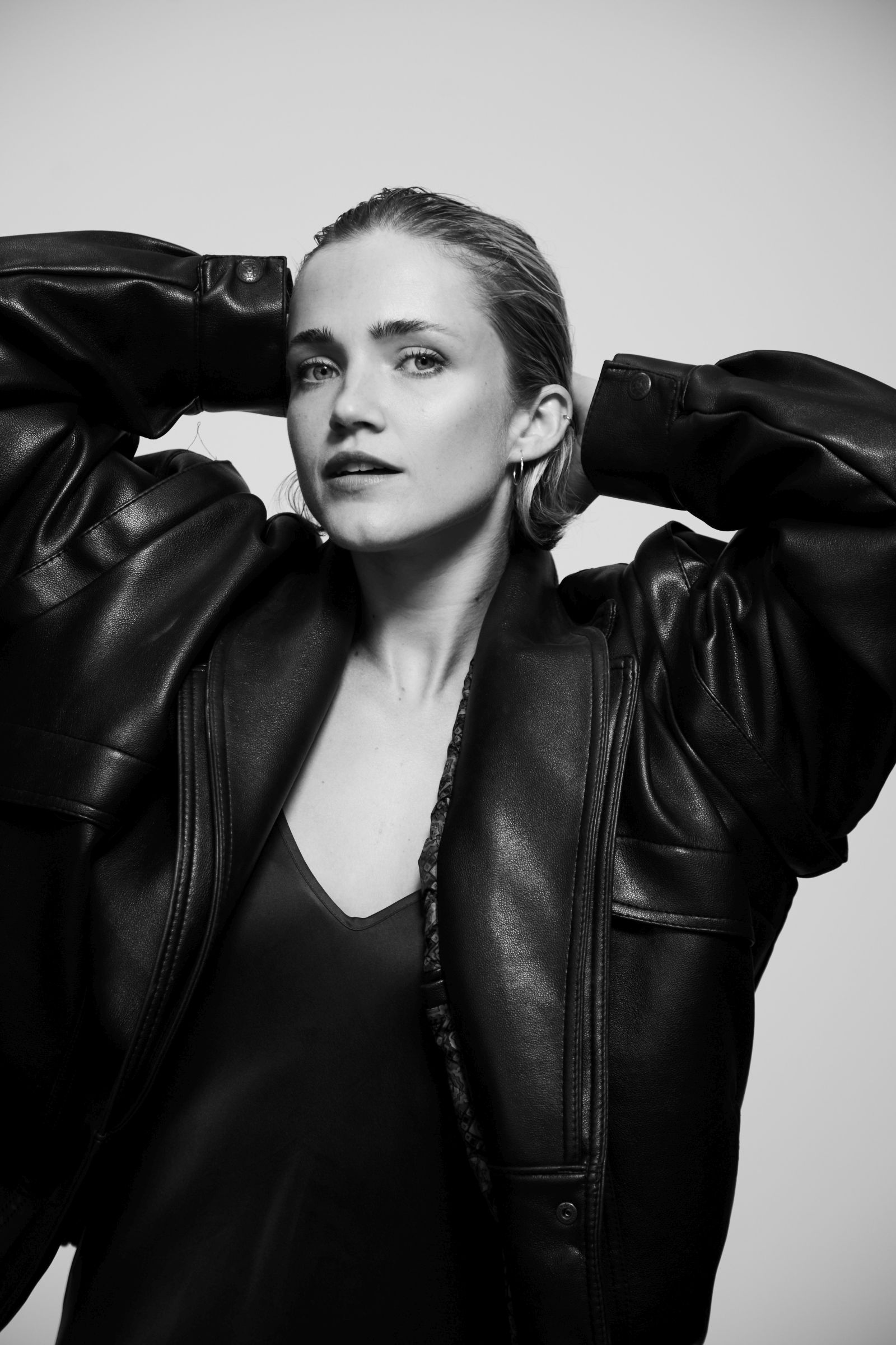 Black-and-white editorial portrait of a person in an oversized leather jacket, hands behind head.
