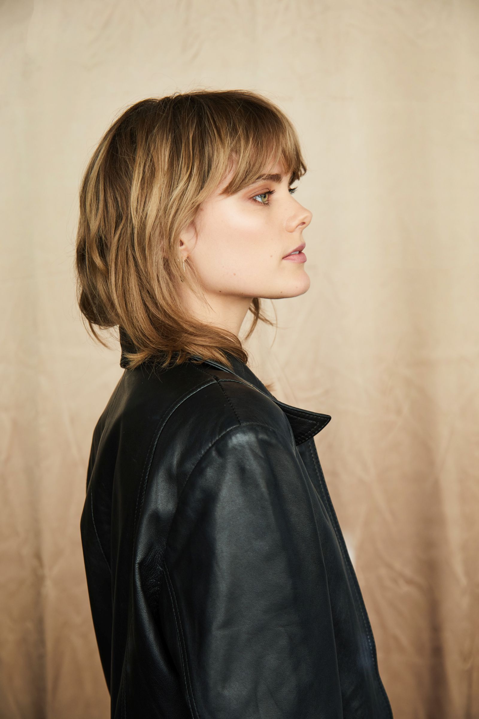 Side-profile studio headshot of person in black leather jacket, neutral beige backdrop.