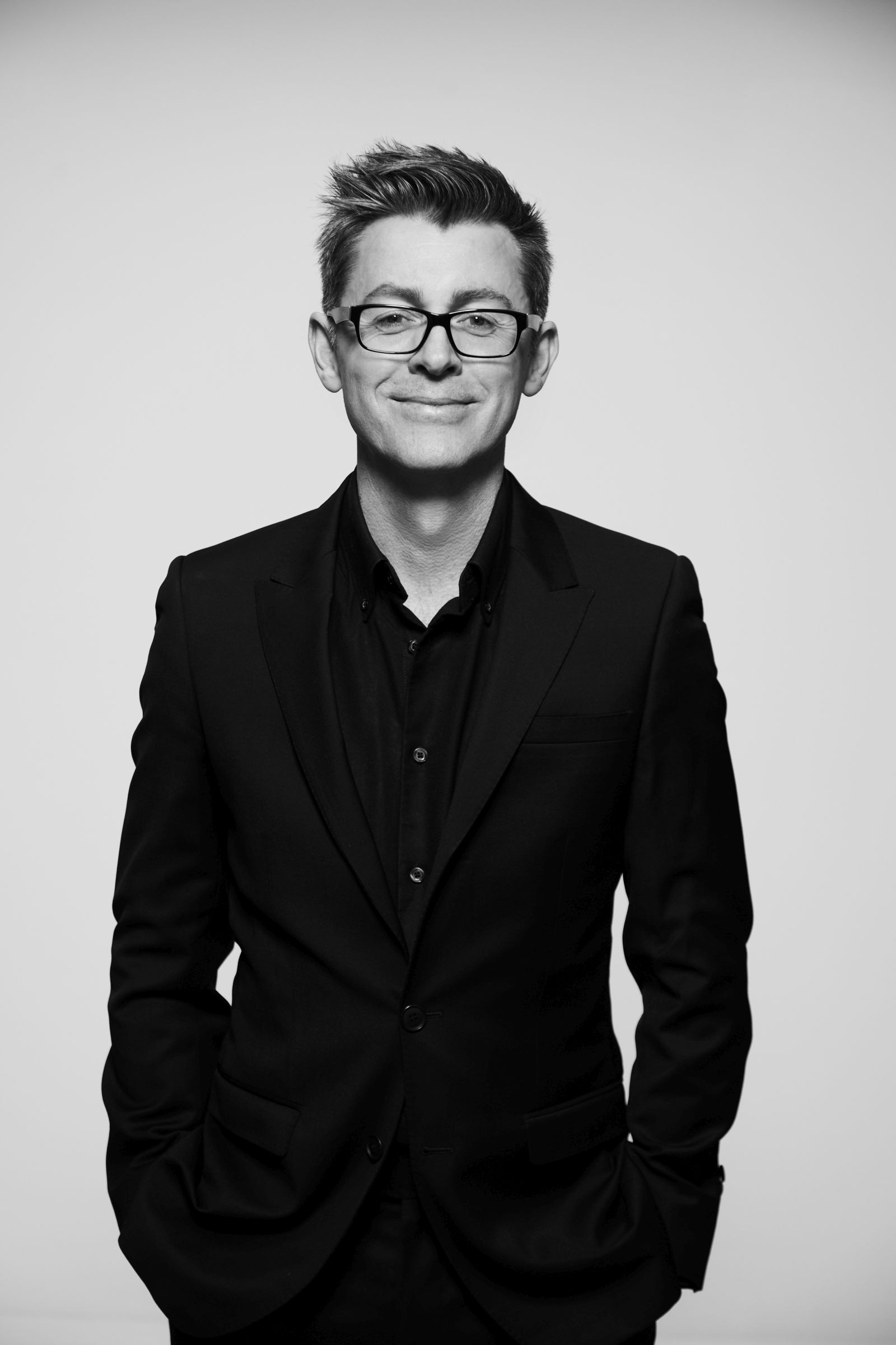 Black-and-white studio portrait of a person in dark suit and glasses, smiling.