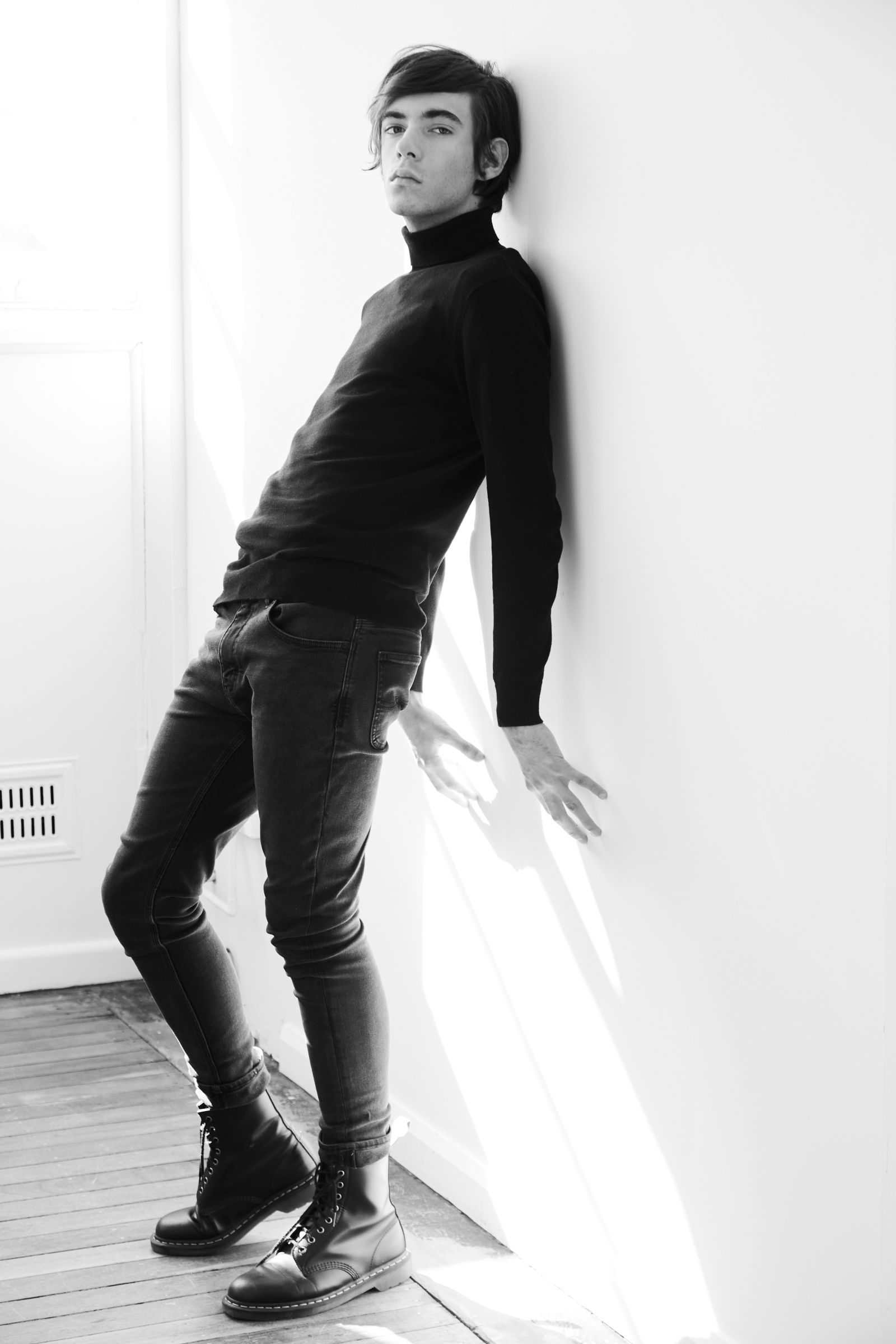 Black-and-white editorial portrait of a person in turtleneck and boots leaning against a sunlit wall.