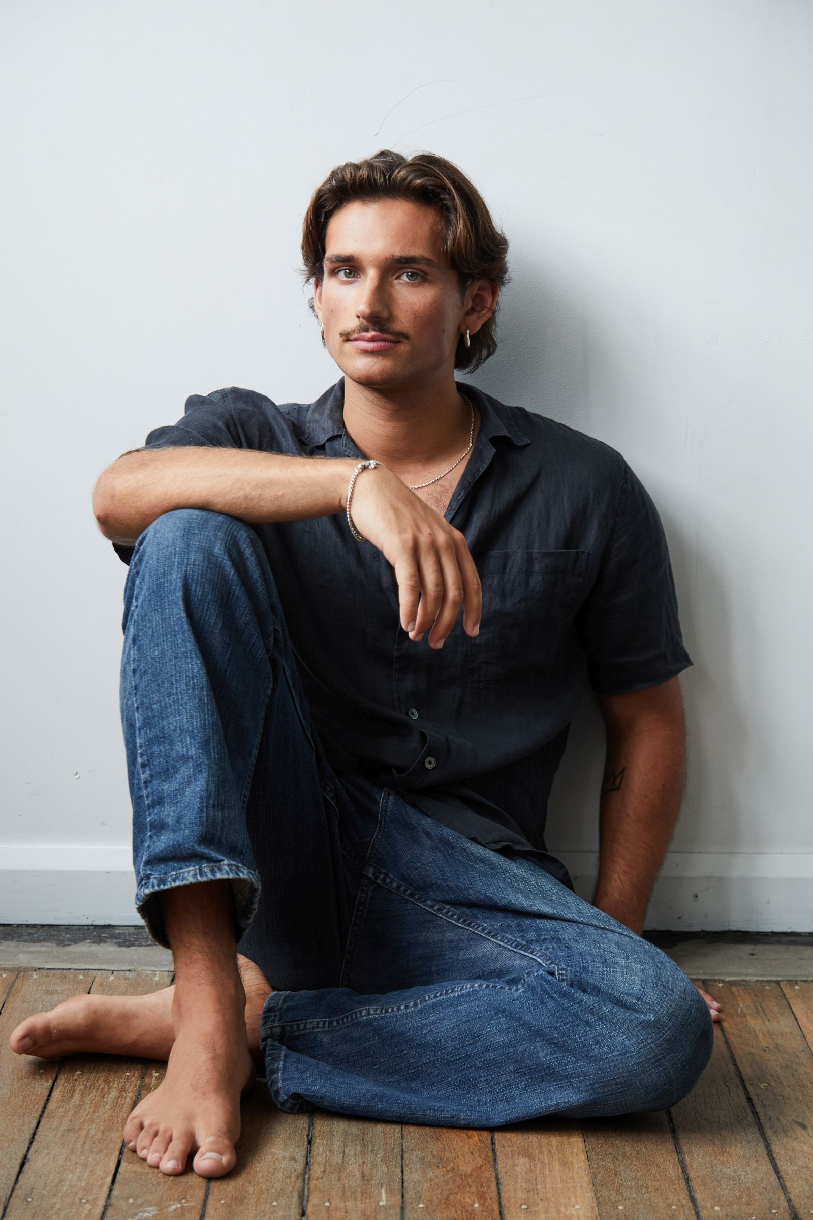 Person seated on floor, wearing denim jeans and a dark shirt, posed against a white wall.