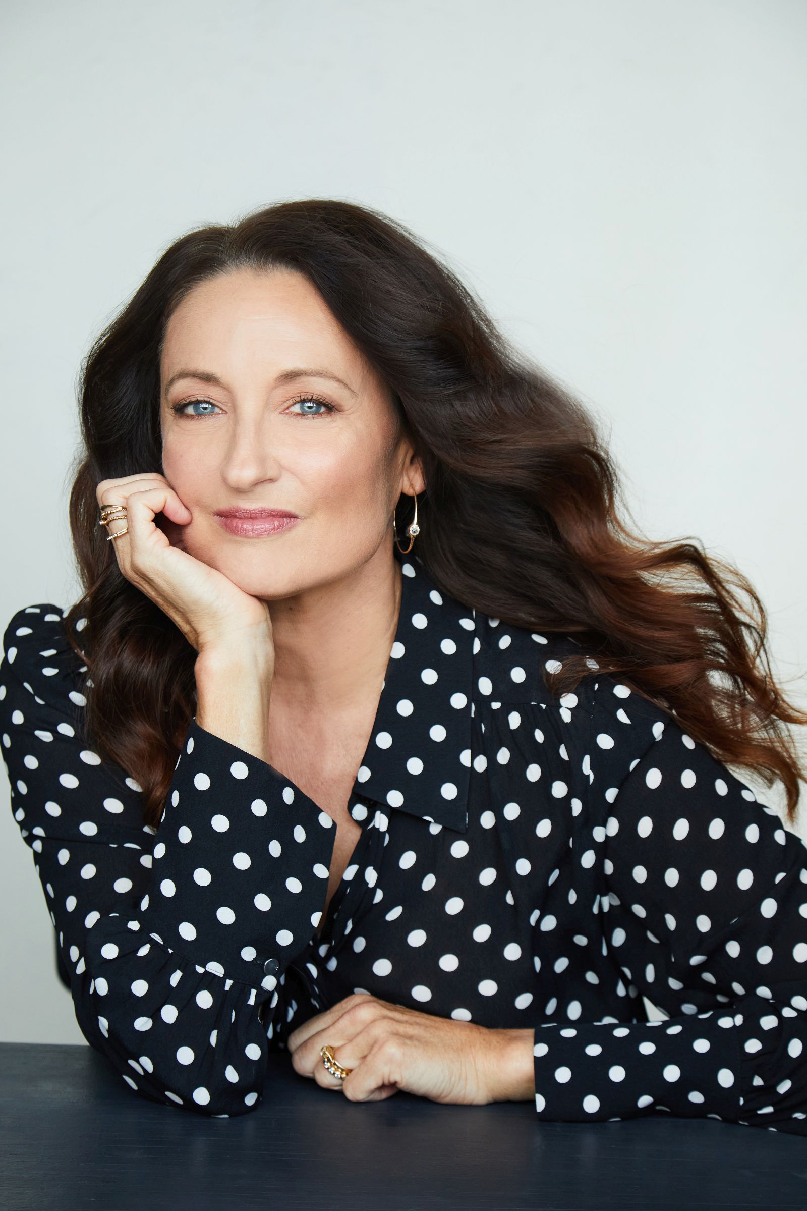 Woman with long dark hair, wearing a polka dot blouse, rests her chin on her hand.