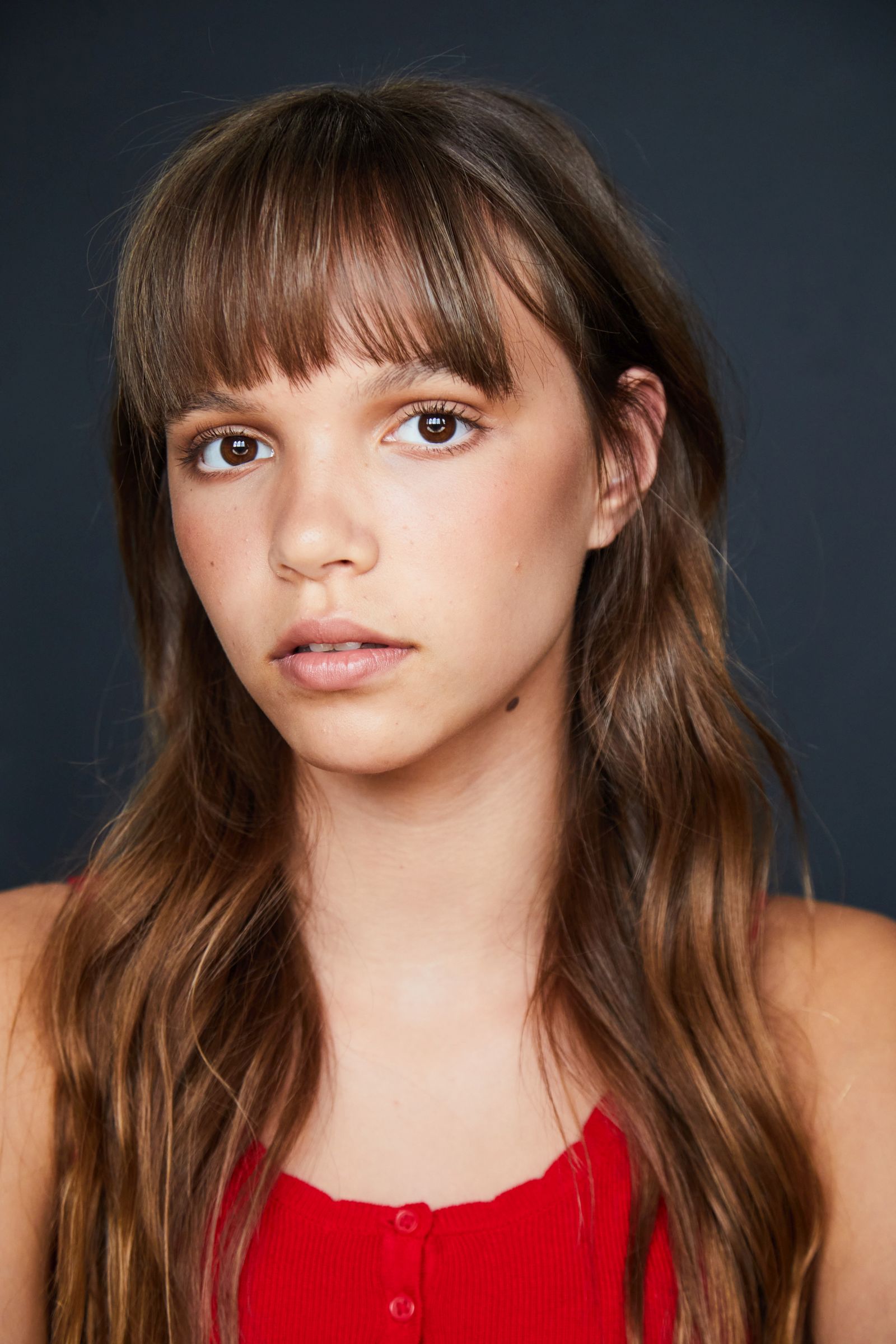 Young person with long hair and bangs, wearing a red top against a dark background.