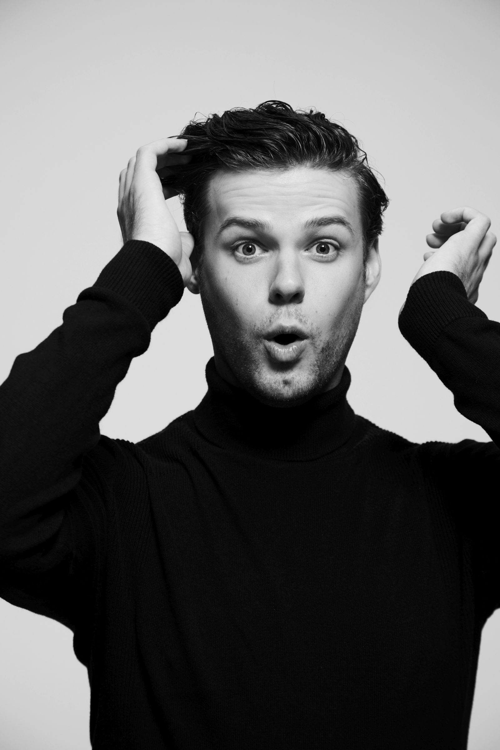Black-and-white studio headshot of man in turtleneck, surprised expression, hands in hair.