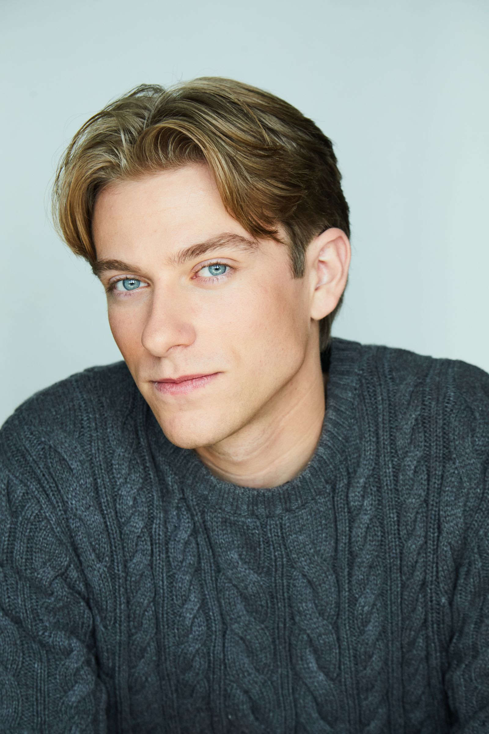 Person with light brown hair wearing a gray cable-knit sweater, against a soft background.