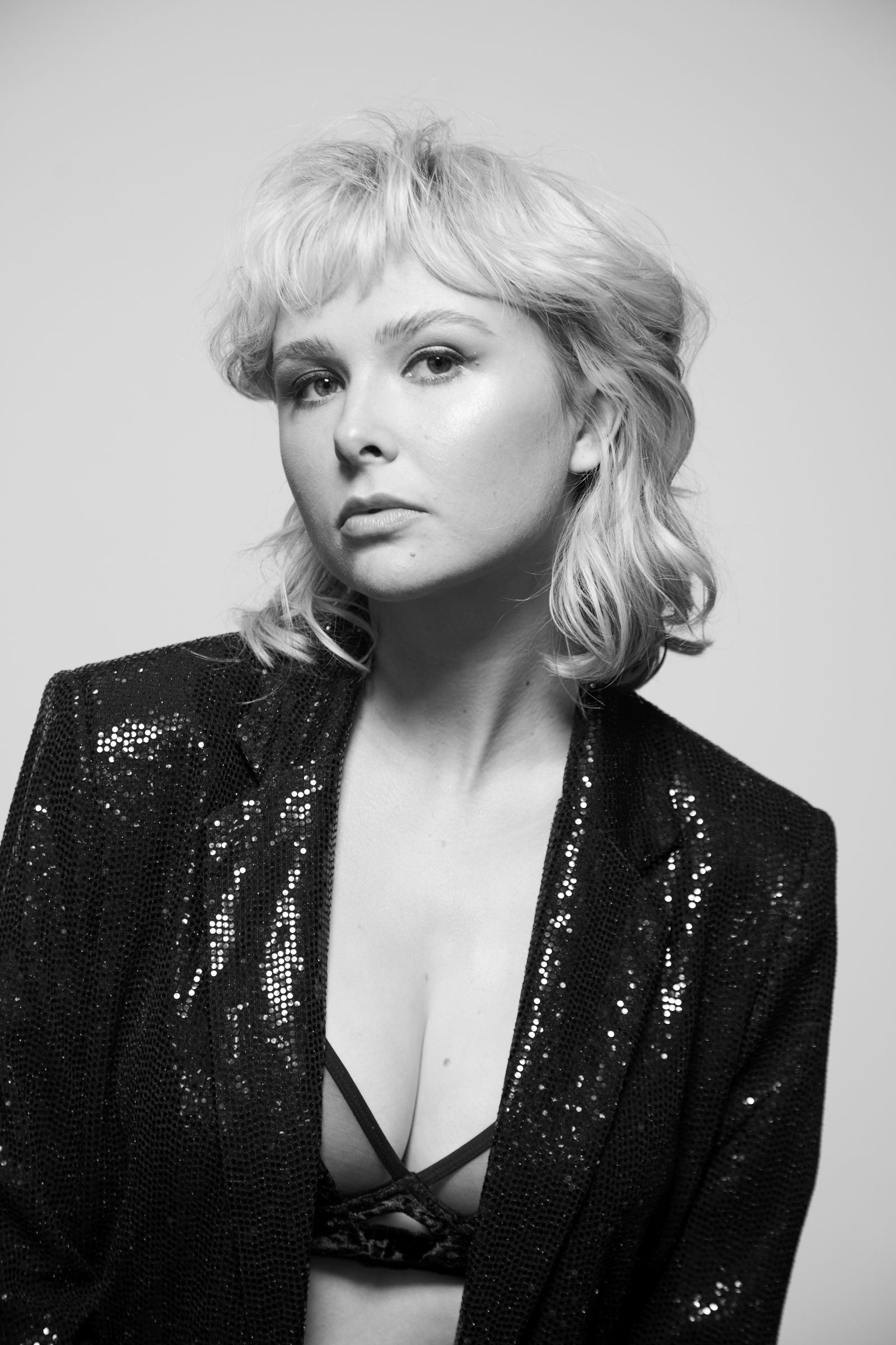 Black-and-white studio portrait of a person with short blonde hair, sequined blazer, strappy bralette.
