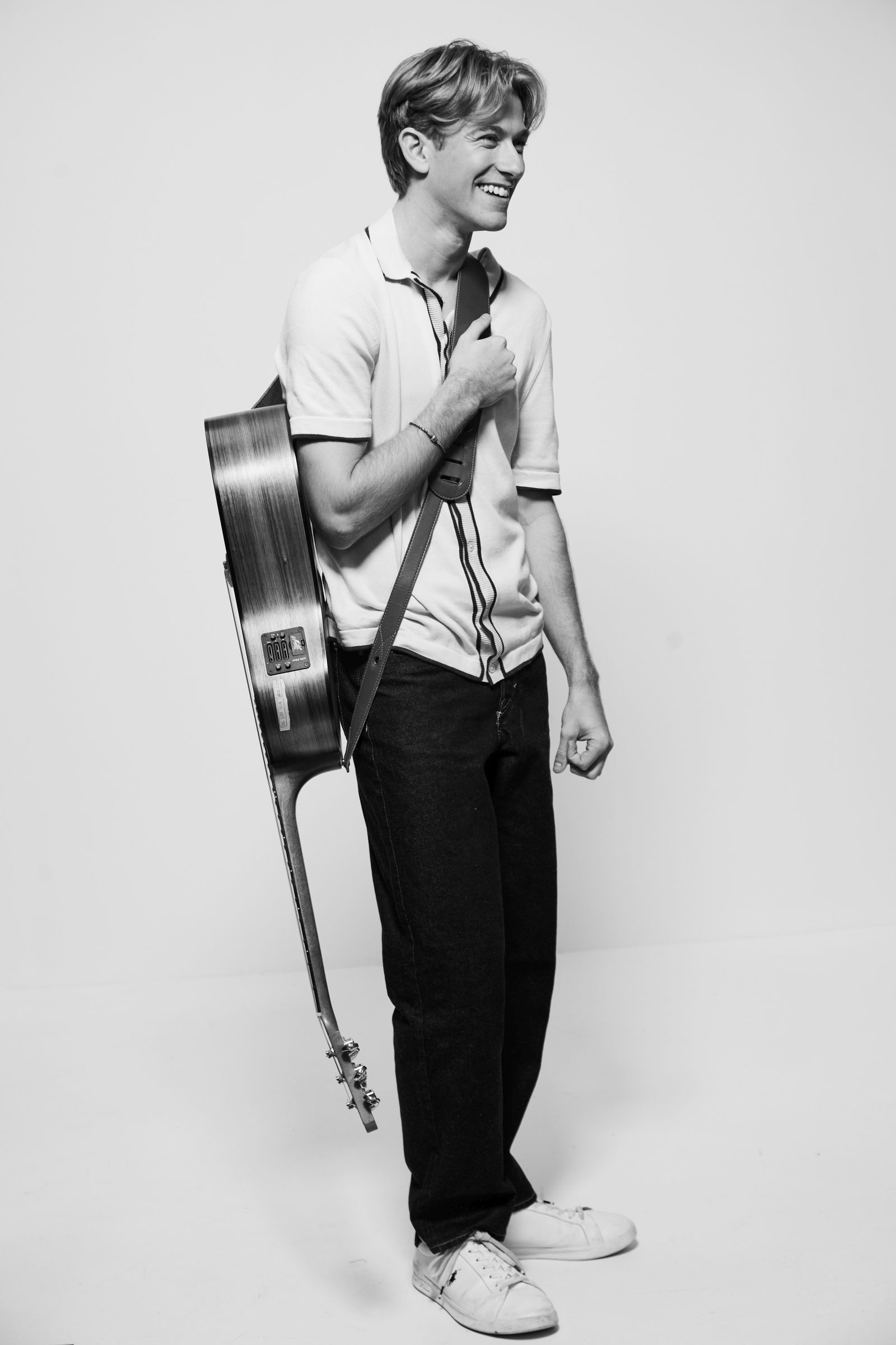 Smiling man carries acoustic guitar on shoulder in black-and-white studio portrait.