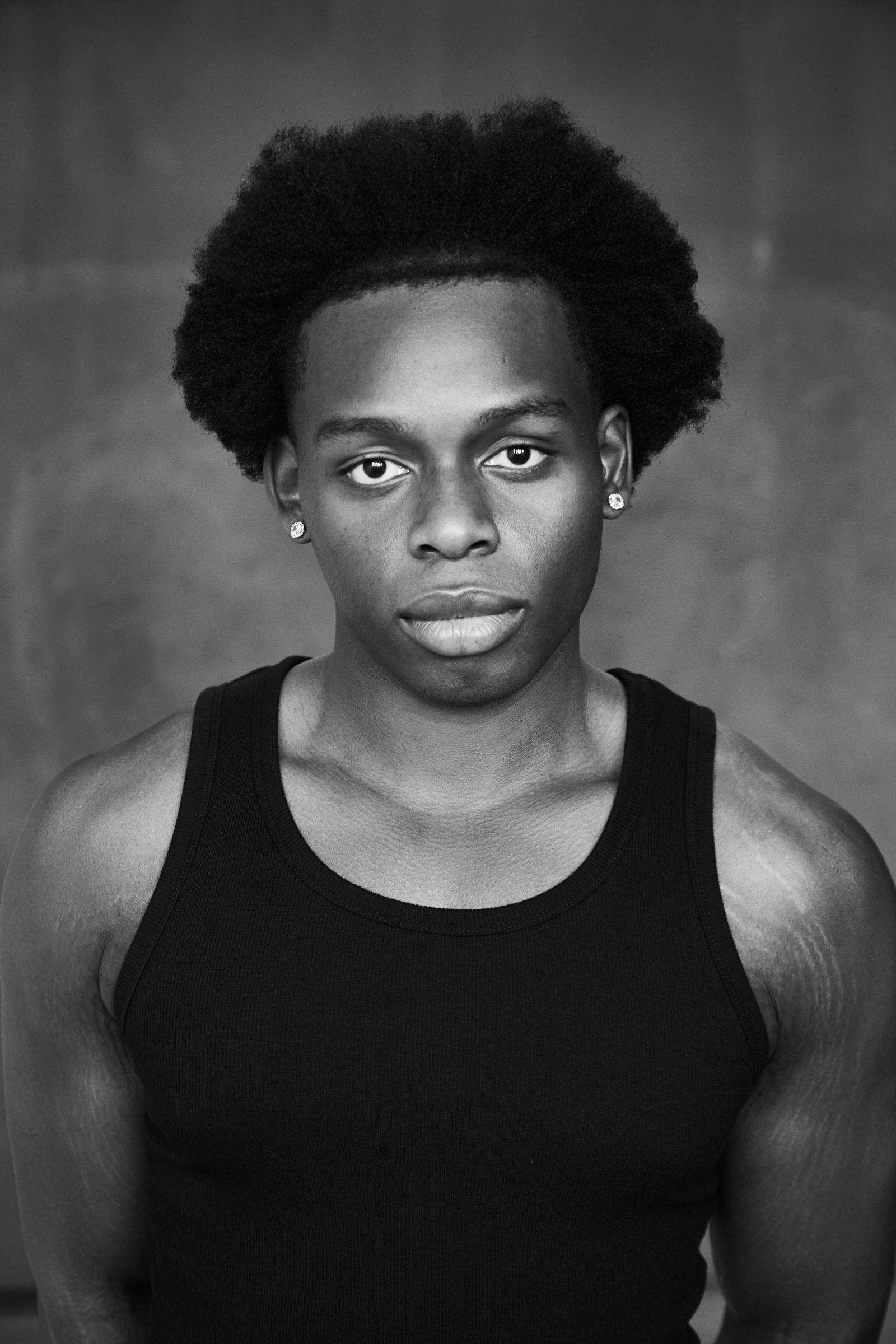 Young man with afro hairstyle in a black tank top against a plain background.
