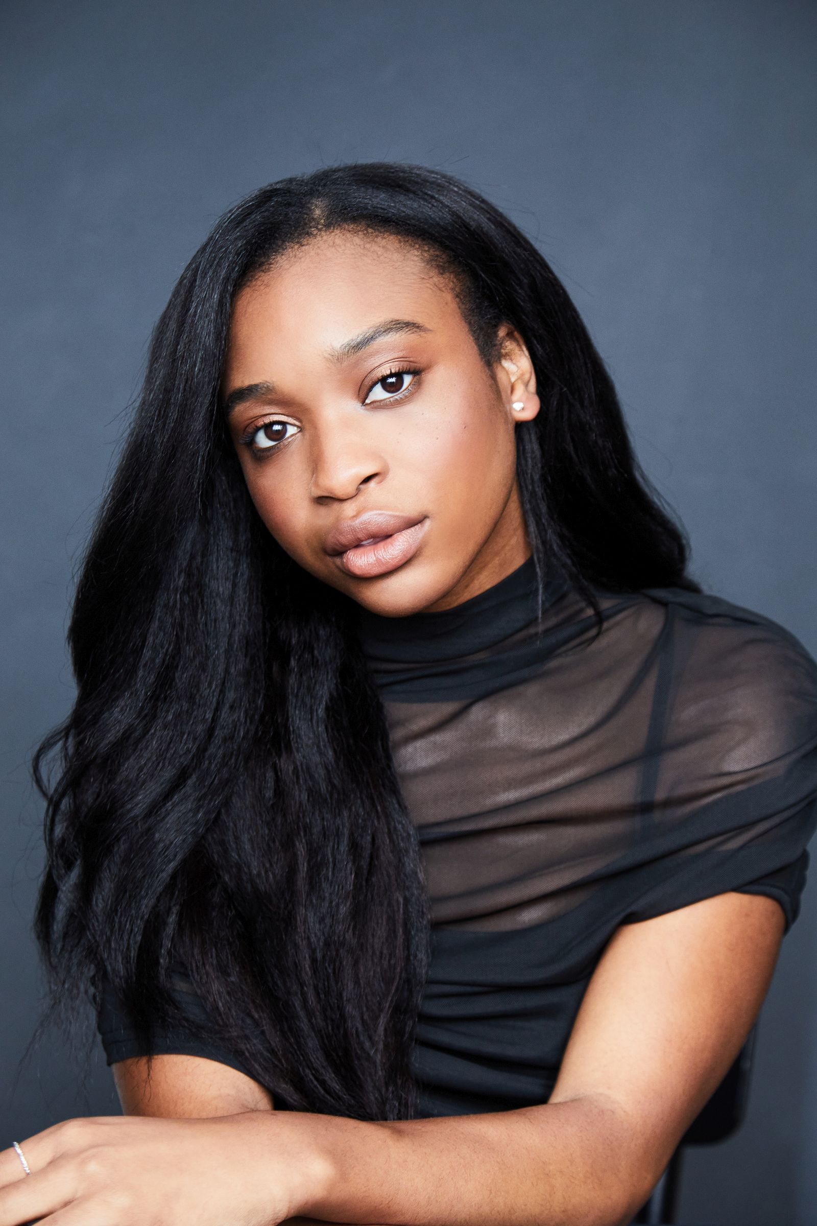 Woman with long dark hair wearing a sheer black top against a dark background.