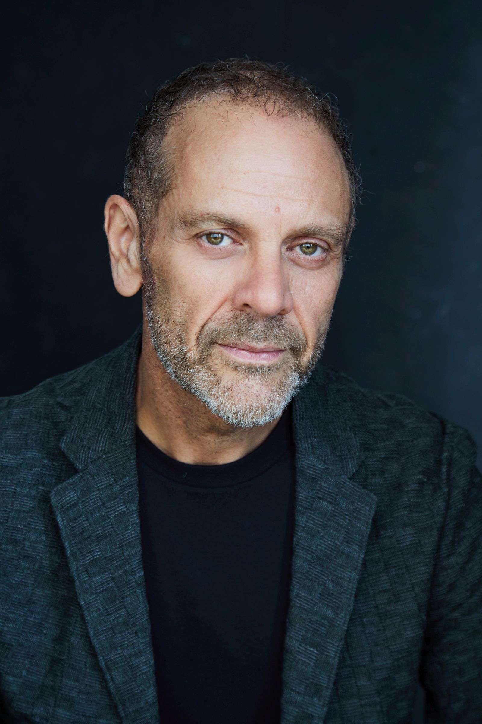Man with short hair and beard wearing a dark jacket against a dark background.