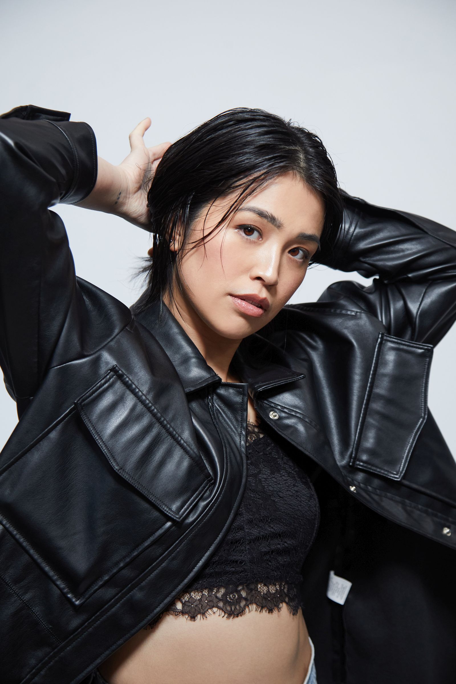 Studio editorial portrait of model in black leather jacket and lace top, hands behind head.