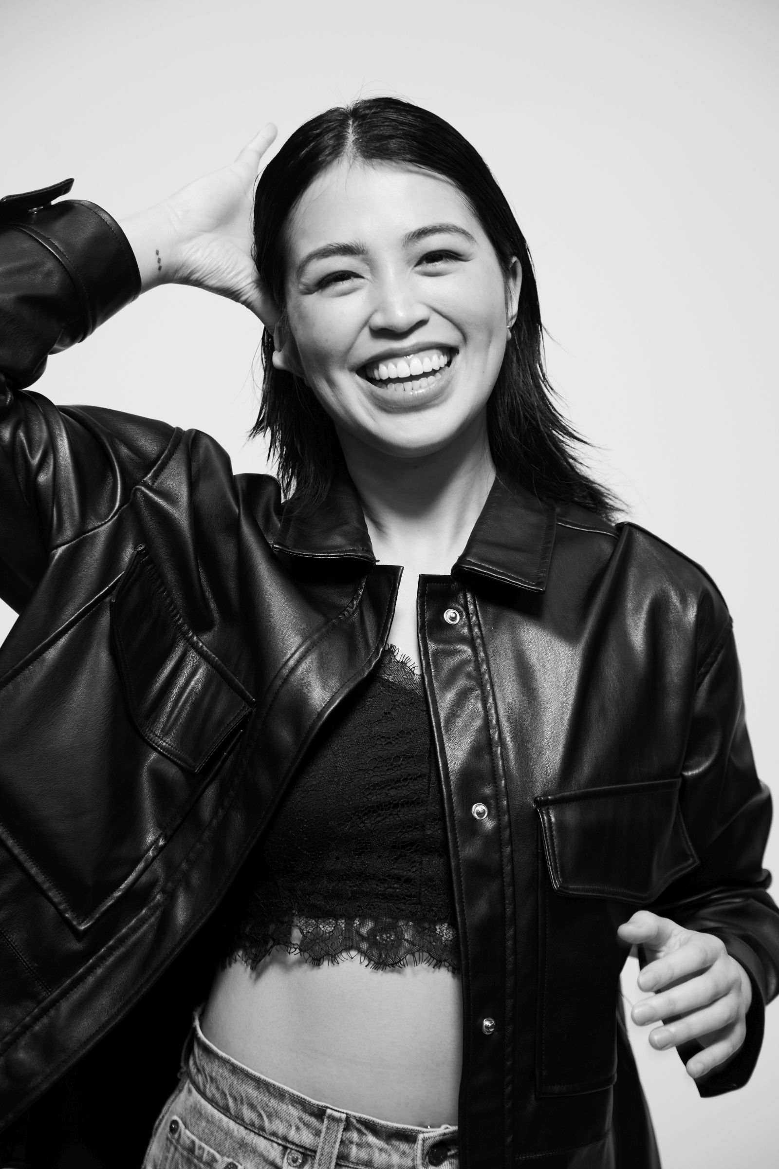 Black-and-white editorial studio portrait of smiling person in leather jacket and lace top, hand raised.