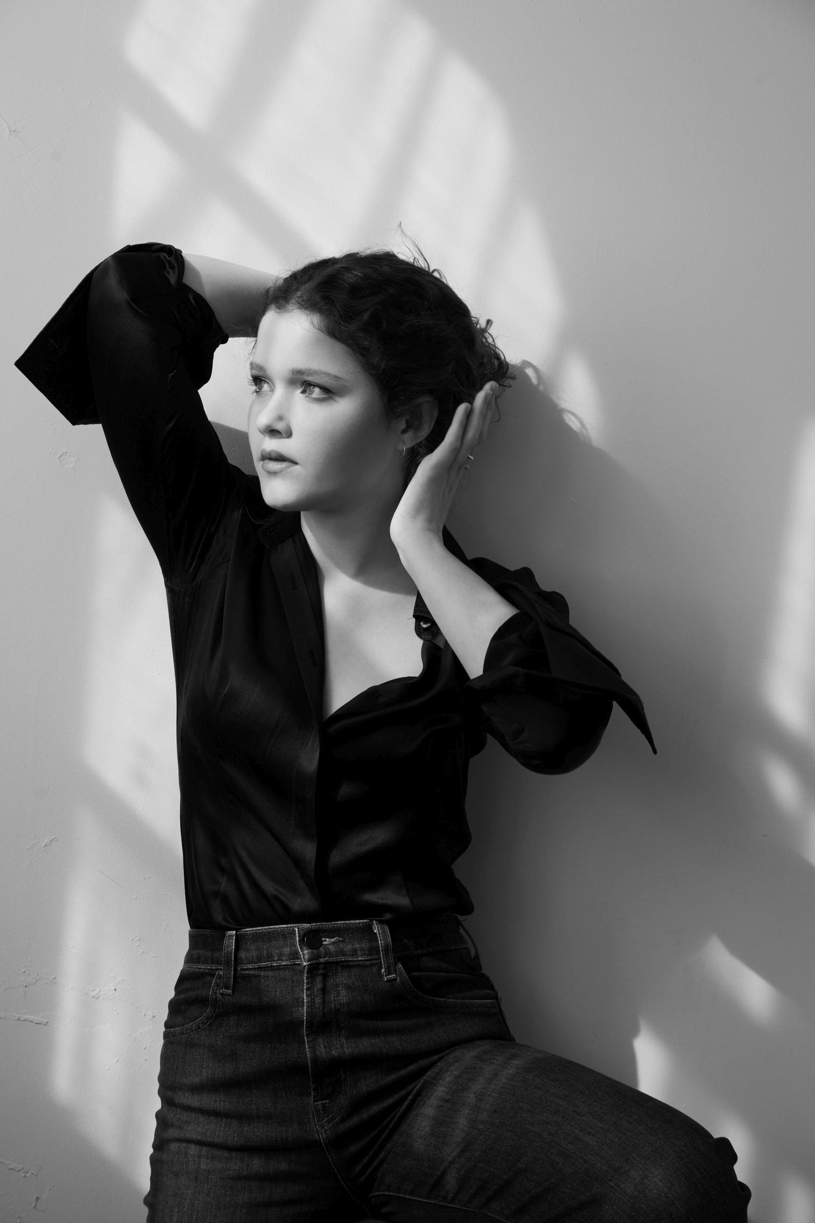 Black-and-white editorial portrait of person in black shirt, hands in hair, window light shadows.