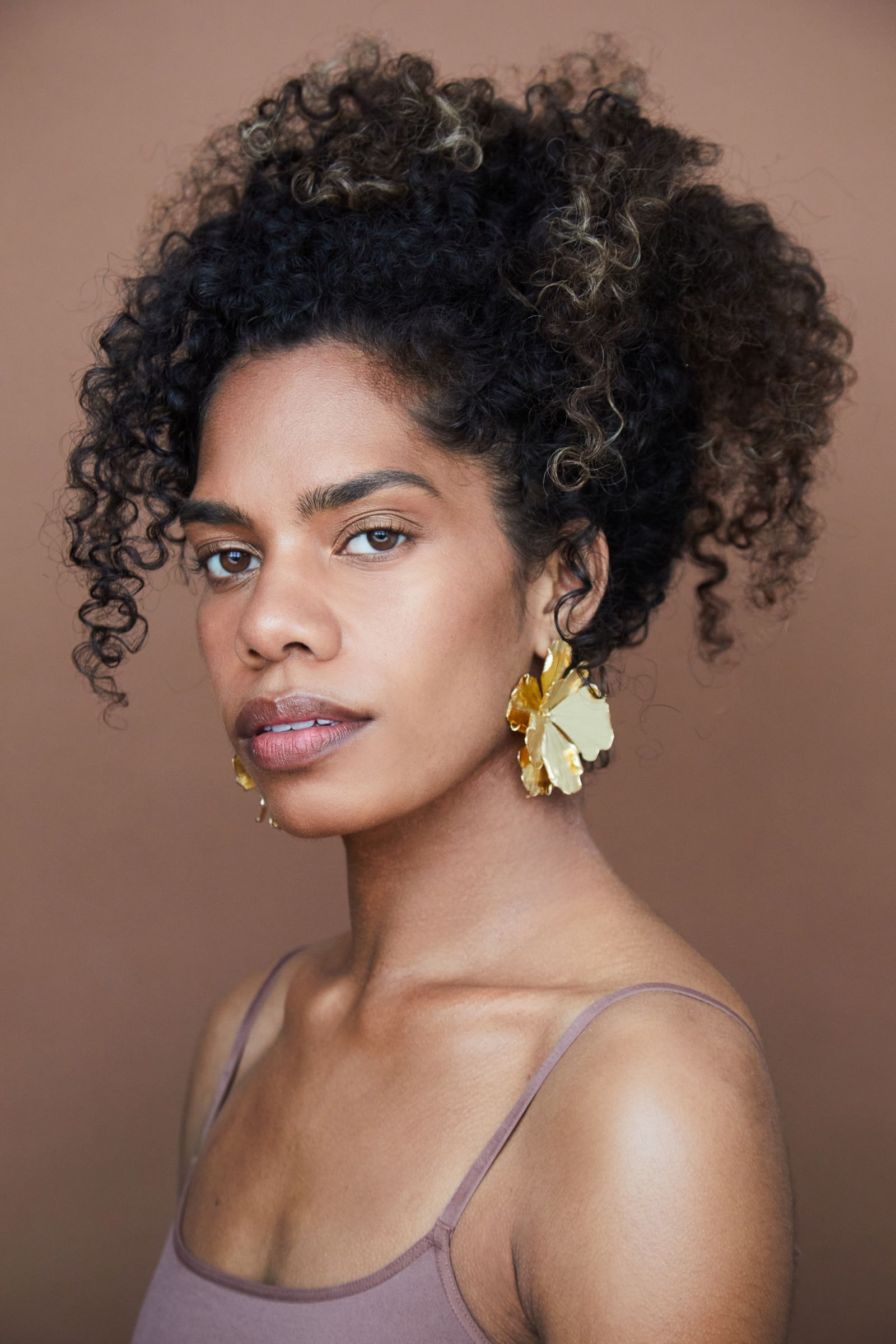 Portrait of person with curly hair and large gold earrings against brown background.