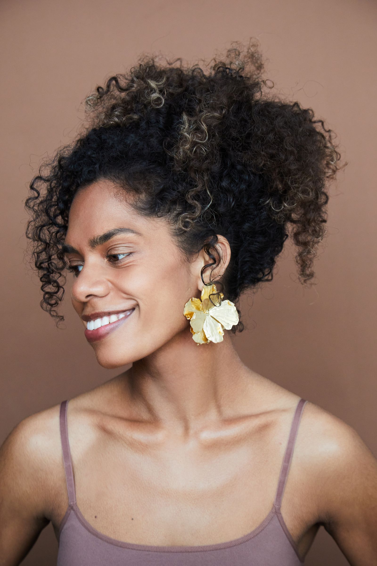 Smiling editorial headshot of person with curly updo, gold floral earrings, brown backdrop.