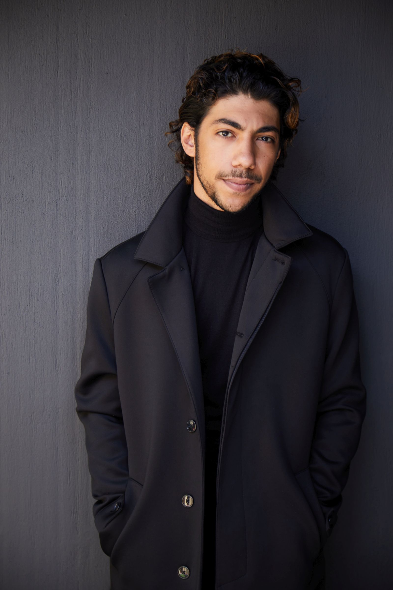 Man in a black coat and turtleneck, leaning against a dark wall.