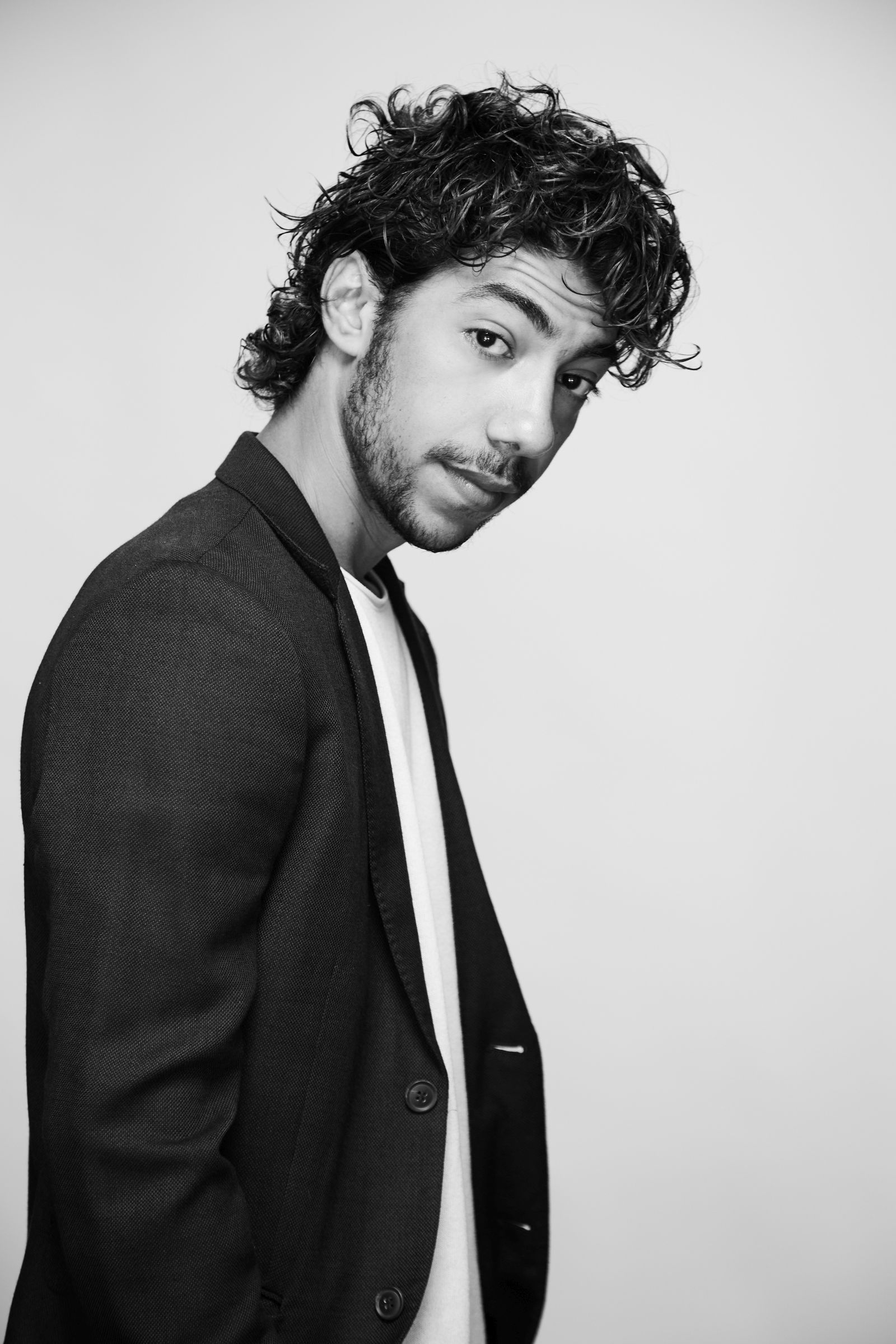 Black-and-white editorial headshot of a curly-haired, bearded person in a blazer.