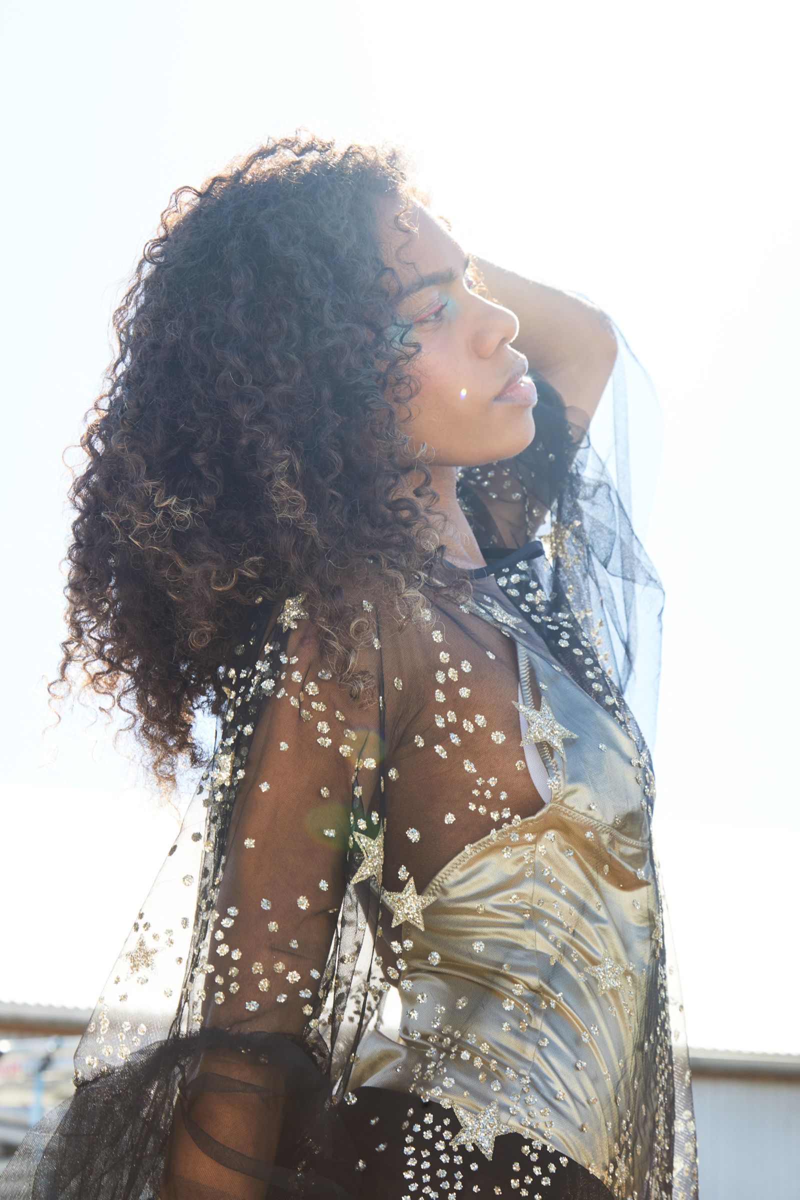 Backlit model with curly hair, arm raised, in starry sheer top and gold corset.