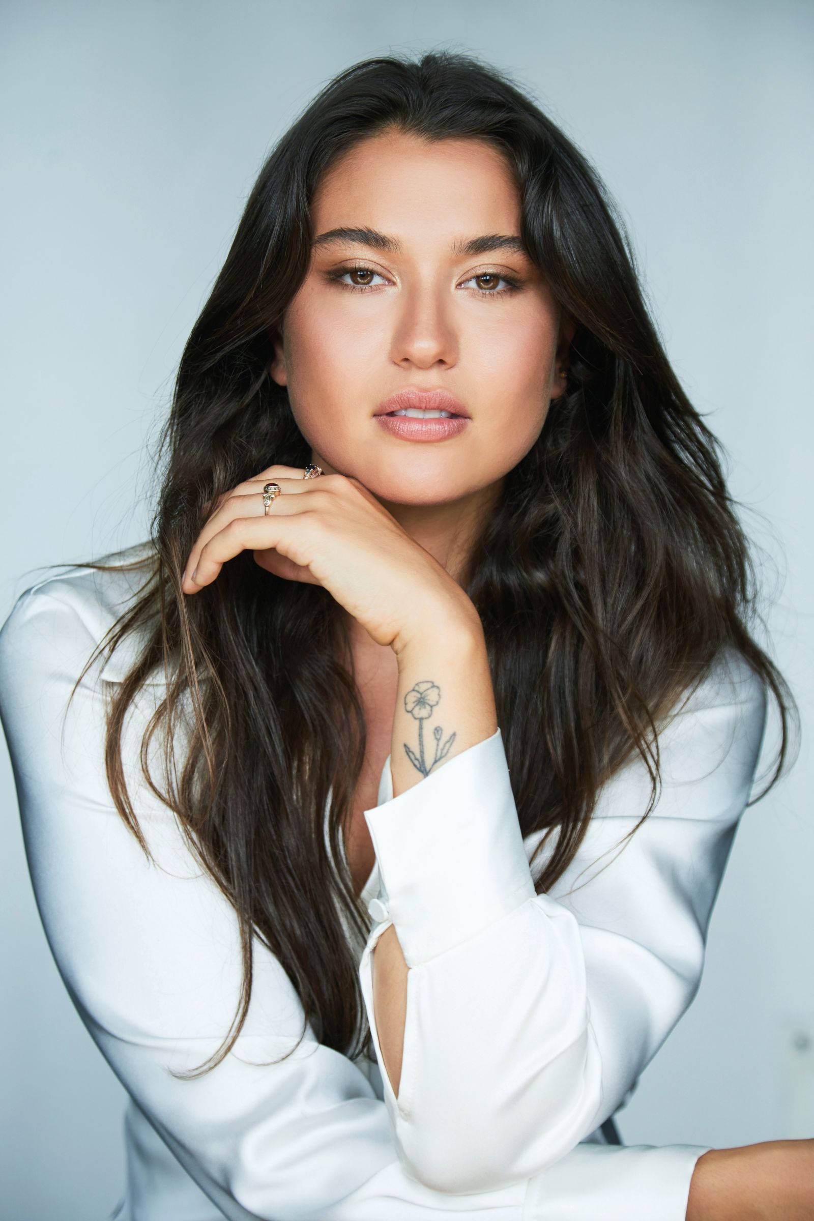 Woman with long dark hair, wearing a white blouse, hand under chin.