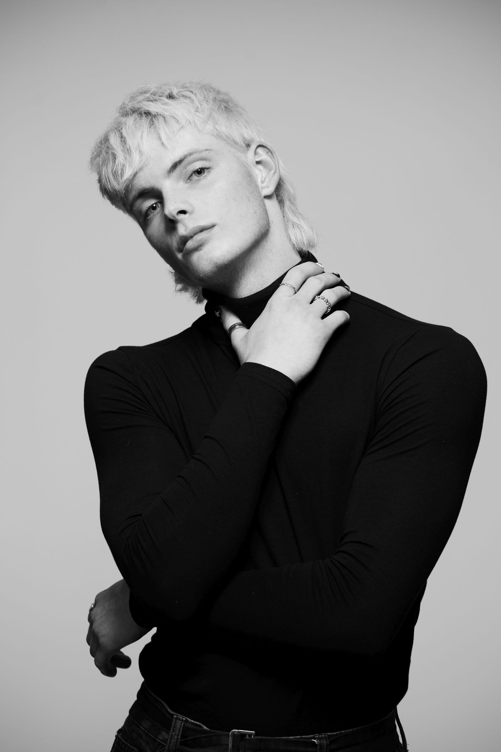 Black-and-white studio editorial portrait of model in black turtleneck, bleached mullet, hand to neck.