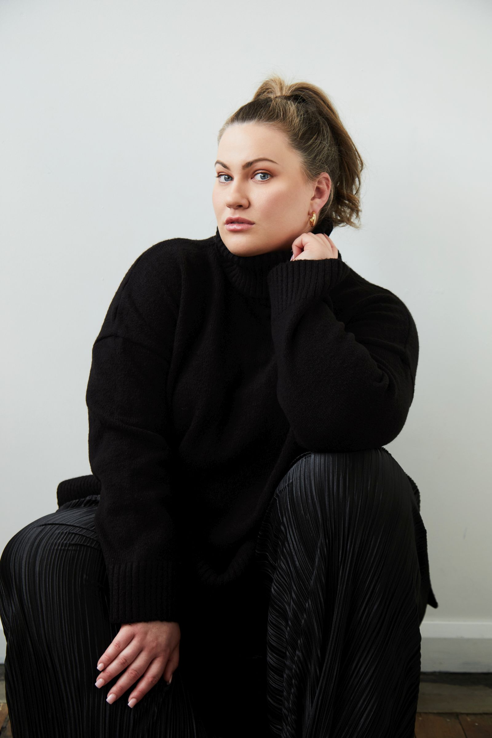 Seated portrait of person in black turtleneck and pleated pants, hand to chin, ponytail, gold hoops.