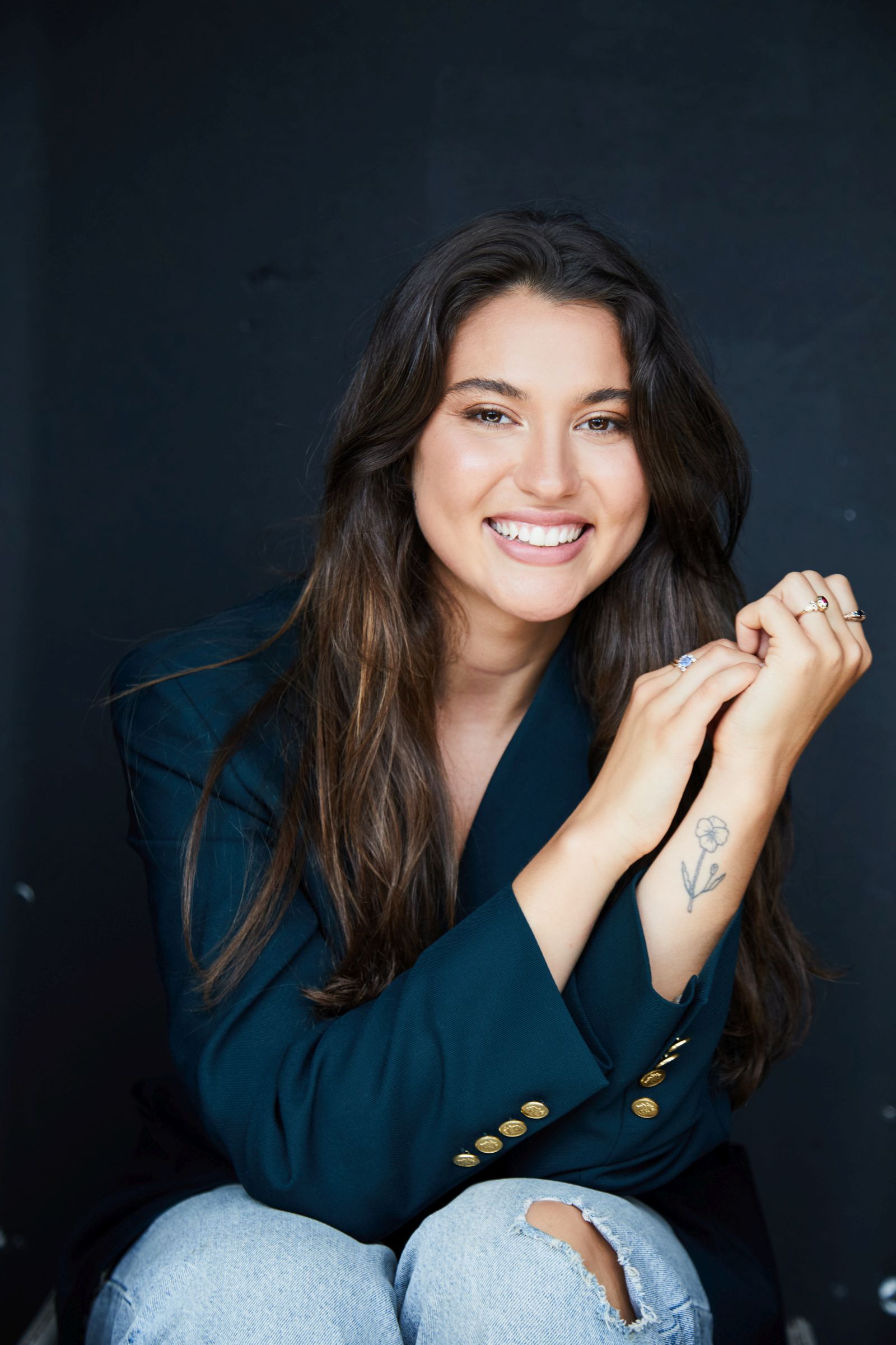 Smiling person with long hair, wearing a blue blazer, floral tattoo on forearm.