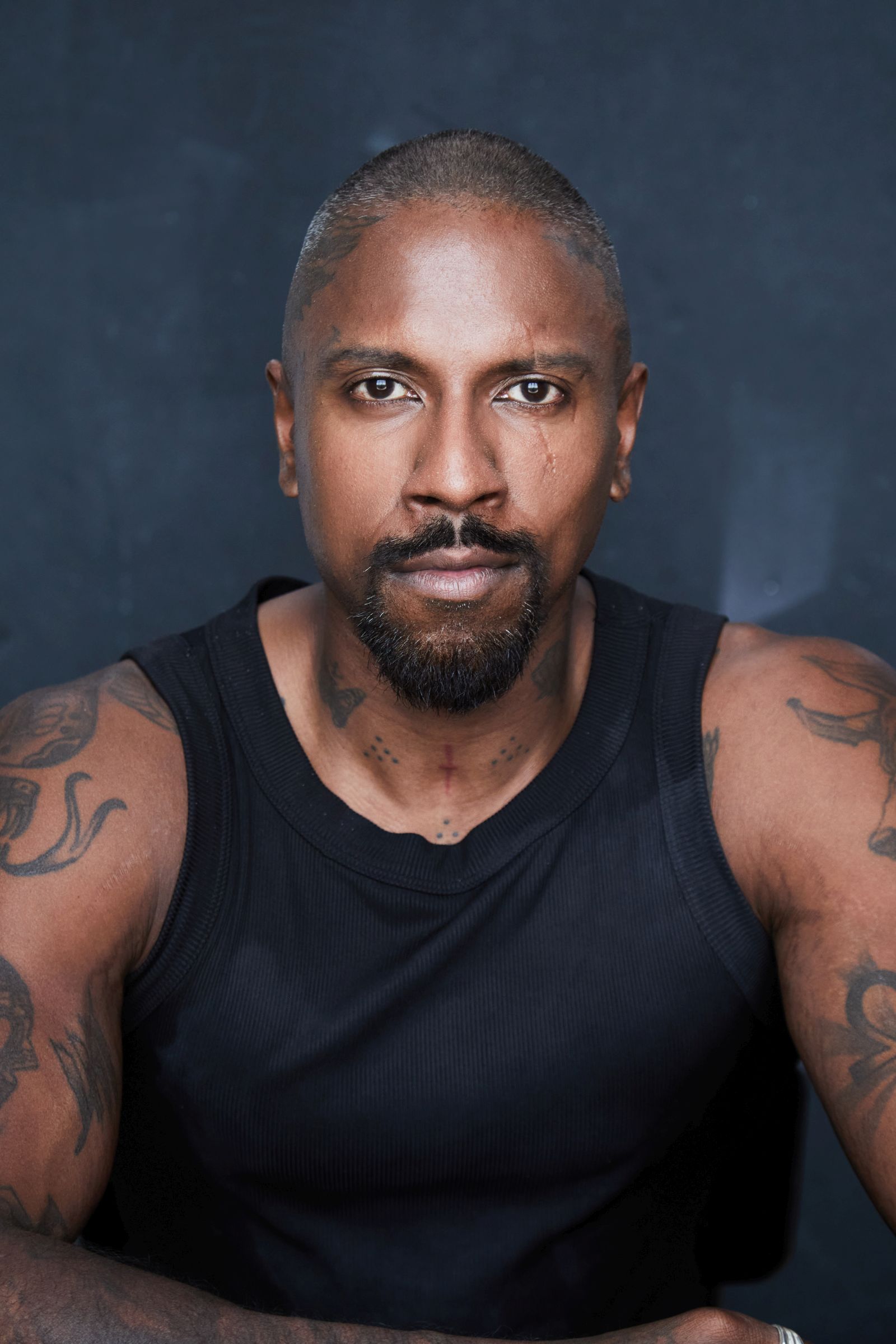 Man with tattoos wearing a black sleeveless shirt, looking directly at the camera.