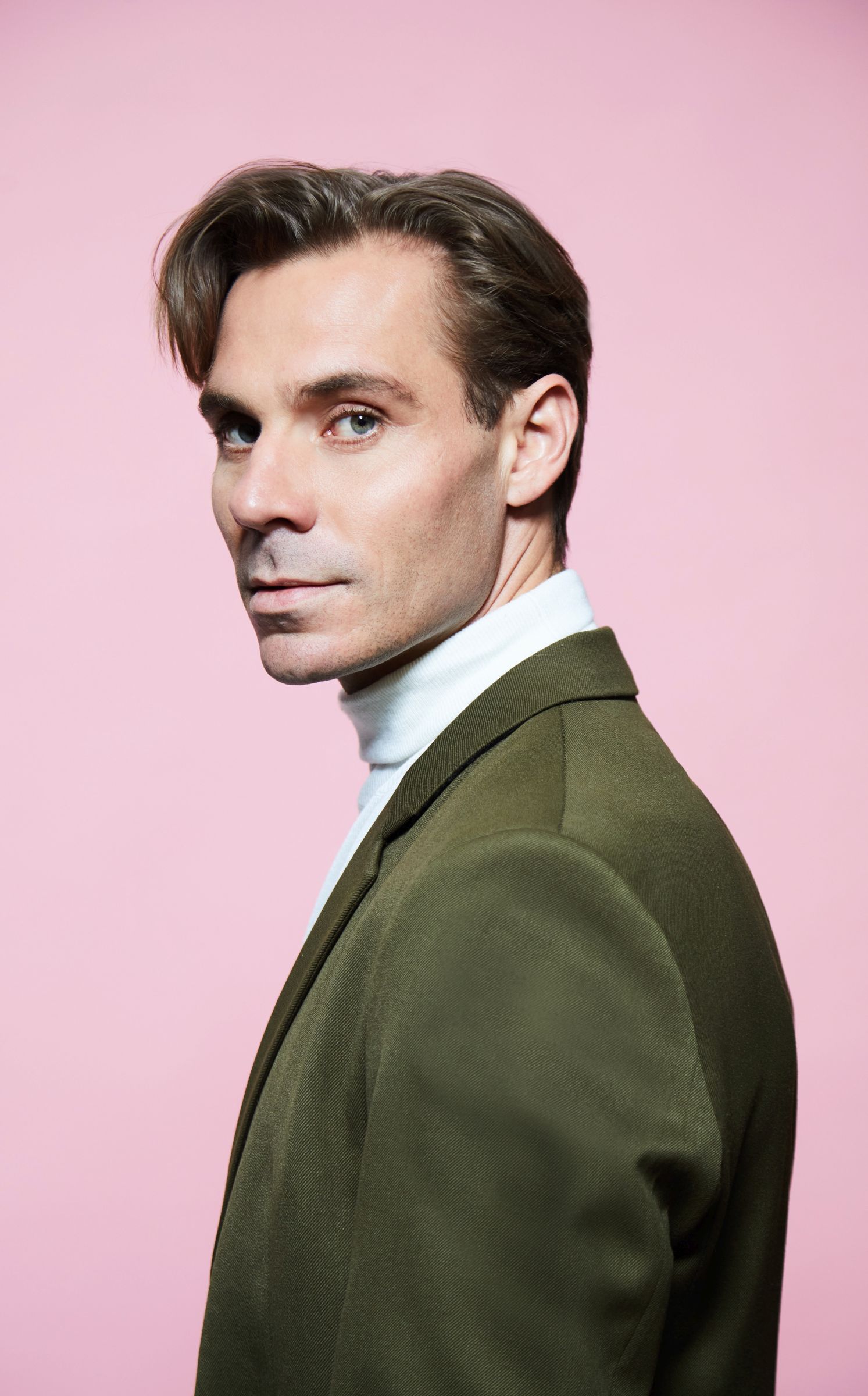Man in olive suit and white turtleneck against pink background.