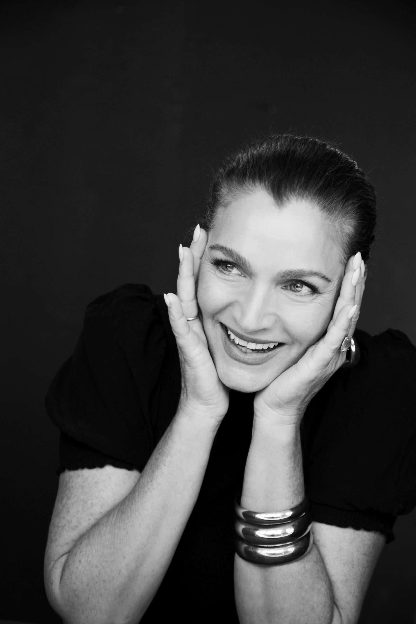 Black-and-white studio portrait of smiling woman cupping her face, dark backdrop, silver bangles.