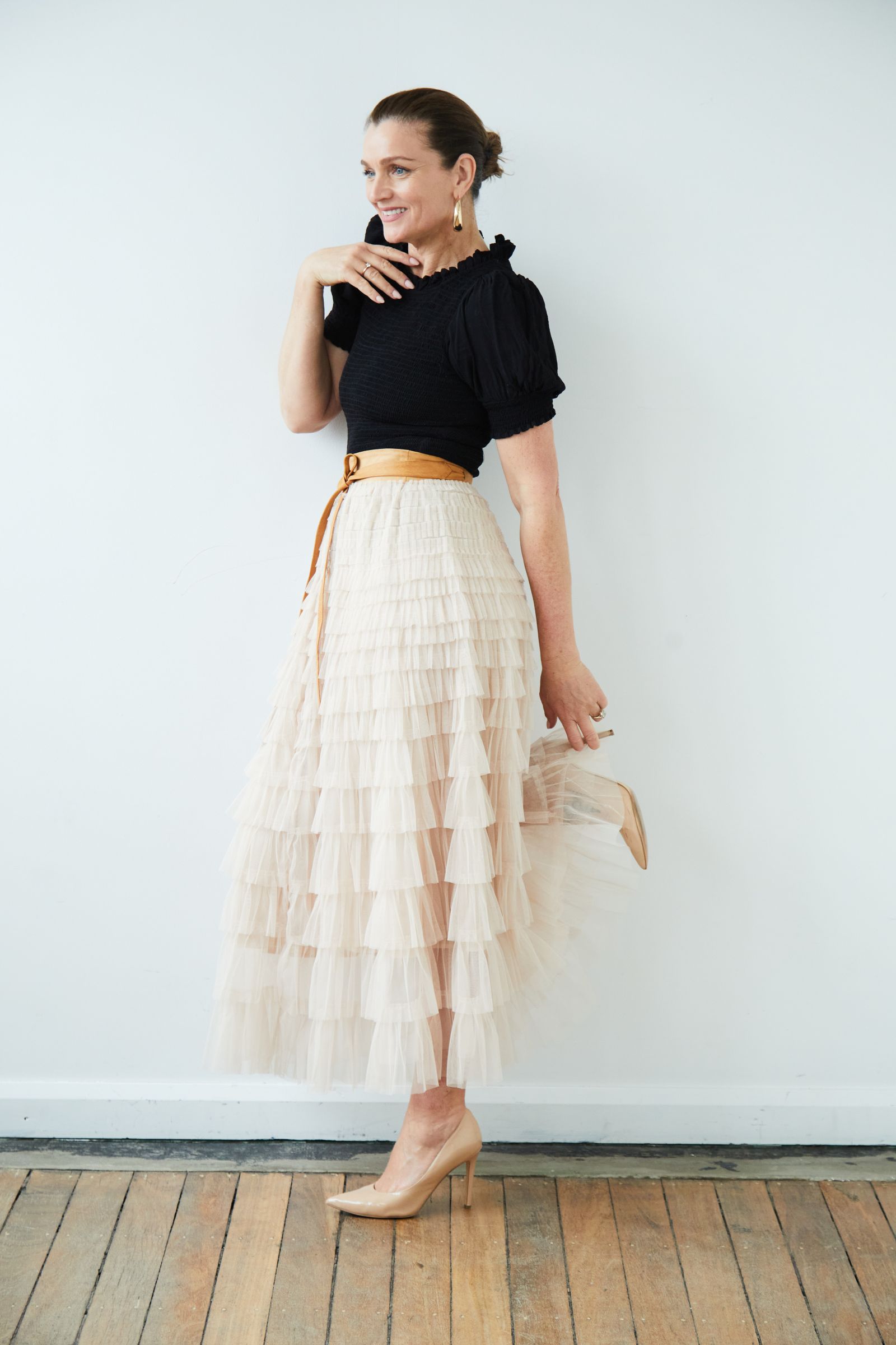 Studio portrait of smiling woman in black top and tiered tulle skirt, lifting one nude heel.